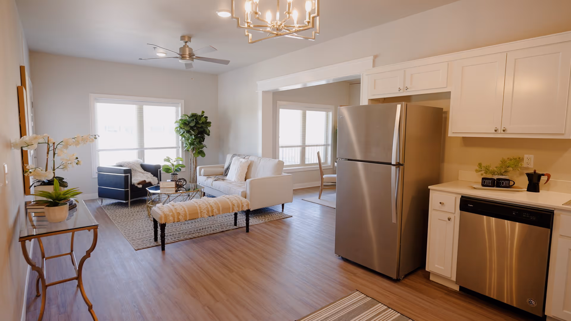 Bright and modern open-concept living space featuring a kitchen area with stainless steel refrigerator and dishwasher, white cabinetry, and a countertop with coffee mugs and a coffee maker. Adjacent living room includes a white sofa, black armchair, glass coffee table, decorative plants, and a patterned rug. Large windows allow natural light to fill the room, and a ceiling fan and chandelier provide lighting.