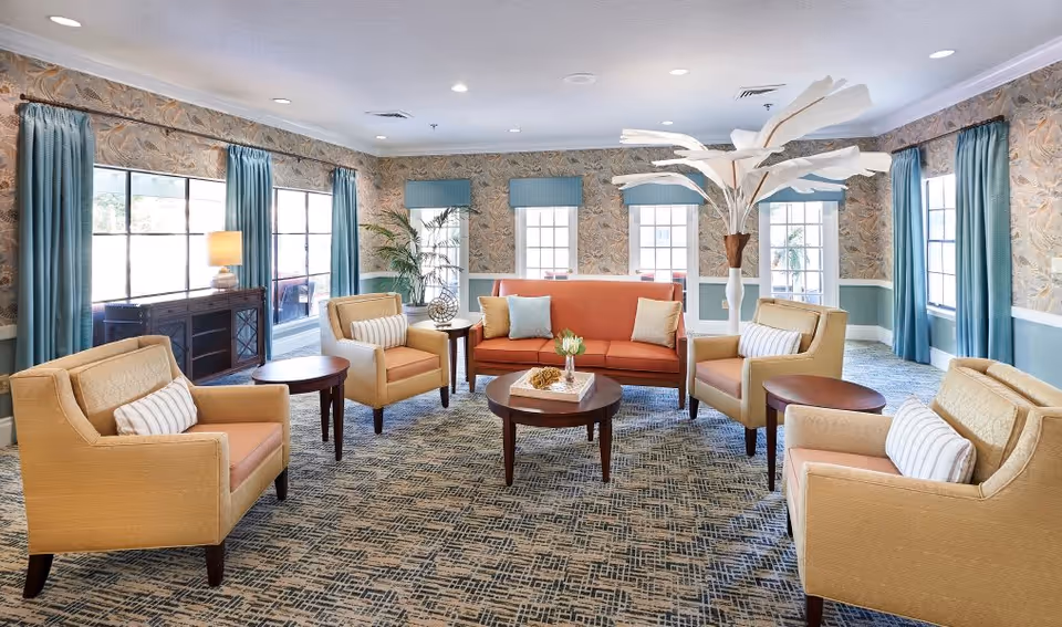 A bright and spacious living room with large windows covered by blue curtains, beige armchairs with striped pillows, an orange sofa with decorative cushions, round wooden coffee tables, a tall white decorative plant, and patterned wallpaper with a blue and beige color scheme.