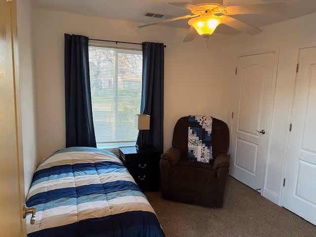 A small bedroom with a single bed covered in a striped blue, white, and beige comforter. Next to the bed is a black nightstand with a lamp. A large window with dark blue curtains is behind the nightstand. To the right is a brown armchair with a blue and white quilt draped over the back. The room has beige carpet and white walls with two closed white doors. A ceiling fan with a light fixture is on the ceiling.
