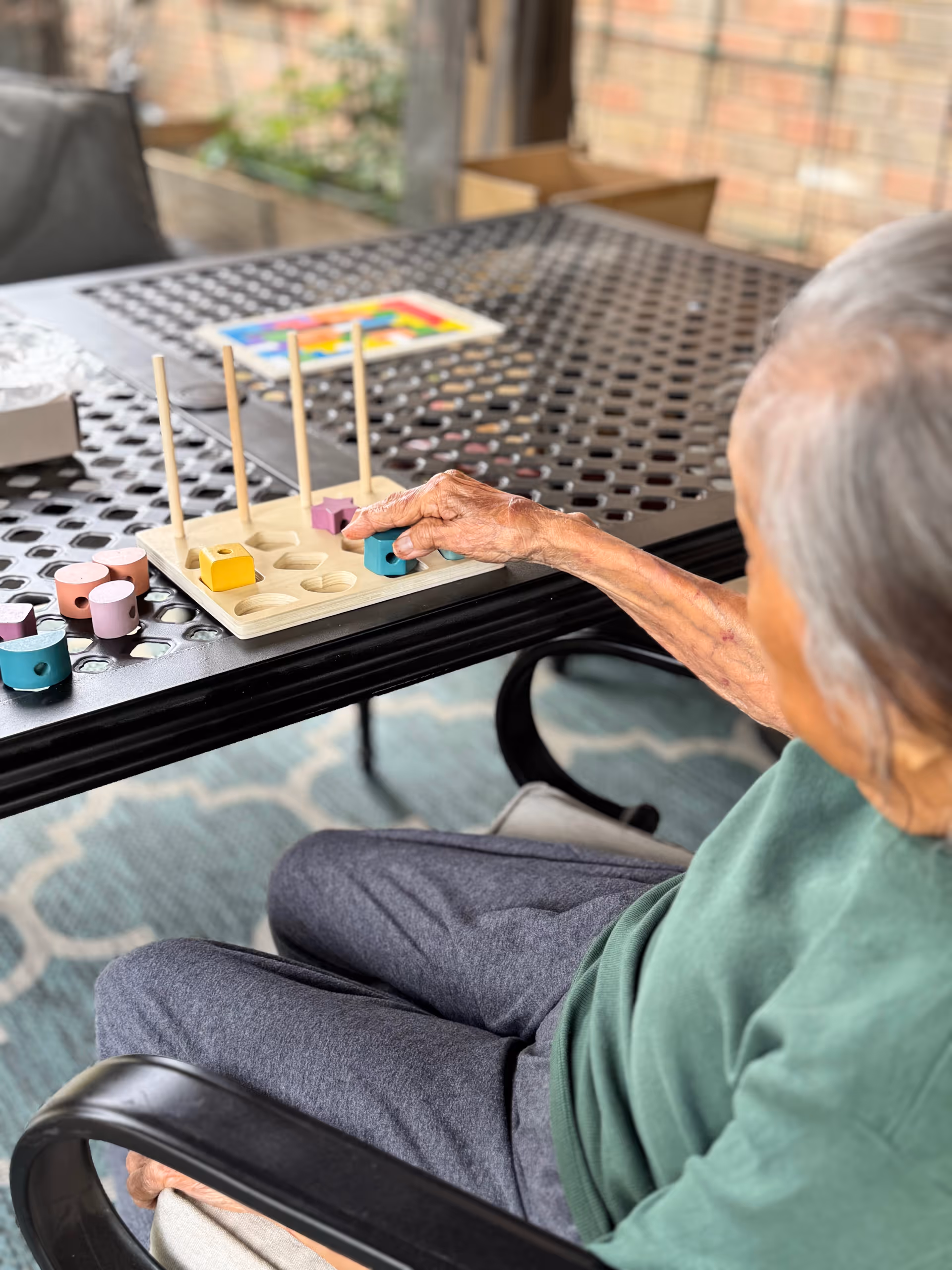 An elderly person seated at a black metal outdoor table playing with a wooden shape-sorting toy that has colorful geometric pieces and vertical pegs. The person is placing a blue piece onto one of the pegs. The background shows a brick wall and some greenery.
