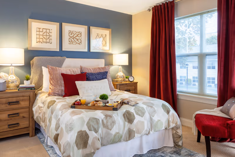 A cozy bedroom with a made bed topped with decorative pillows and a breakfast tray, flanked by wooden nightstands and lamps, framed art above the headboard, and a window with red curtains.