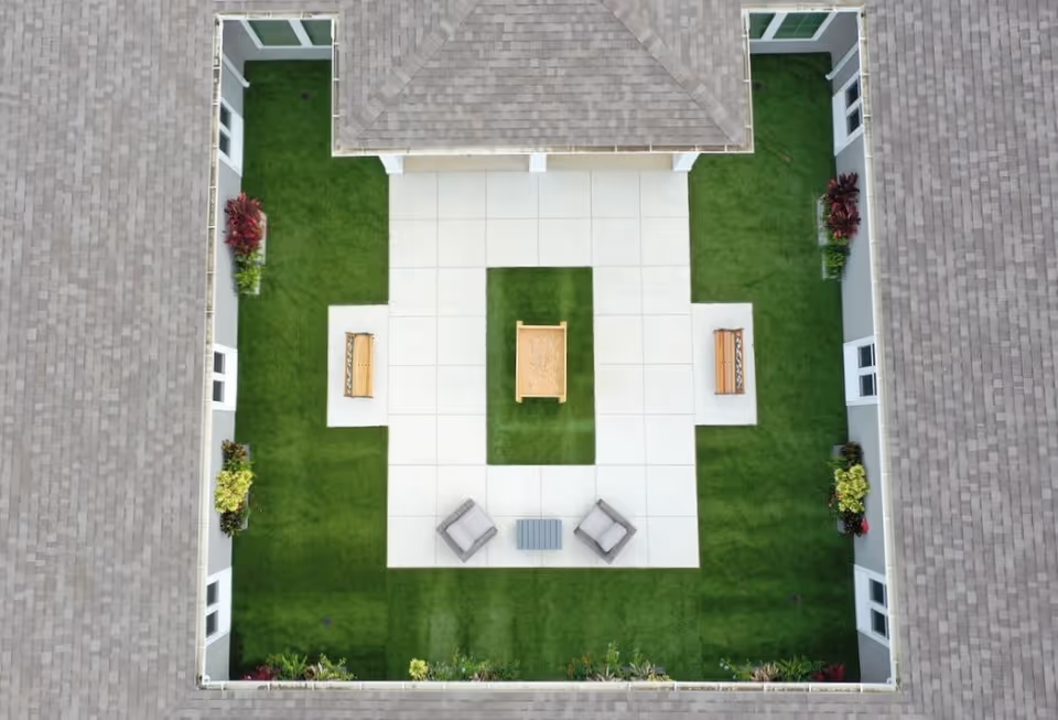 Aerial view of a square interior courtyard with paved walkways, green lawn, benches, chairs and a central planter surrounded by building roofs.
