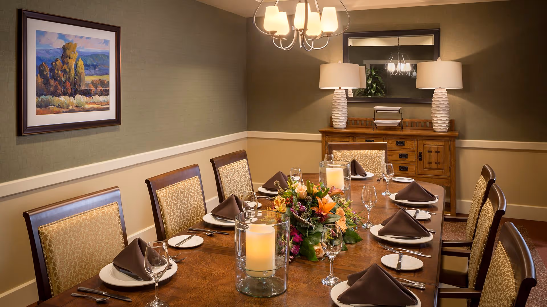 A formal dining room with a wooden table set for eight people. Each place setting includes a white plate, brown folded napkin, wine glass, and silverware. The table is decorated with two large glass candle holders and a colorful floral centerpiece. The room has beige walls with a framed landscape painting on one side and a wooden sideboard with two white lamps and a mirror on the other. A chandelier with multiple lights hangs above the table.
