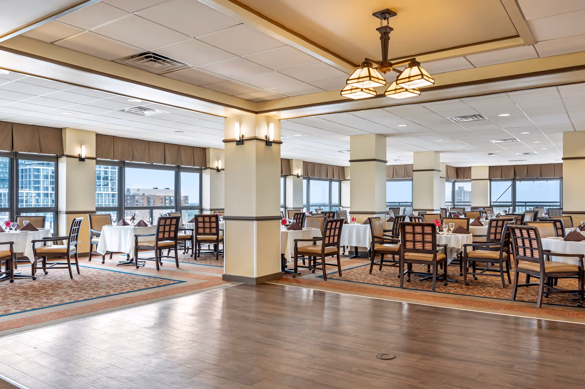 Spacious dining room with multiple tables covered in white tablecloths, each set with brown napkins and glassware. The room has large windows offering a city view, beige walls with columns, and a decorative ceiling light fixture.