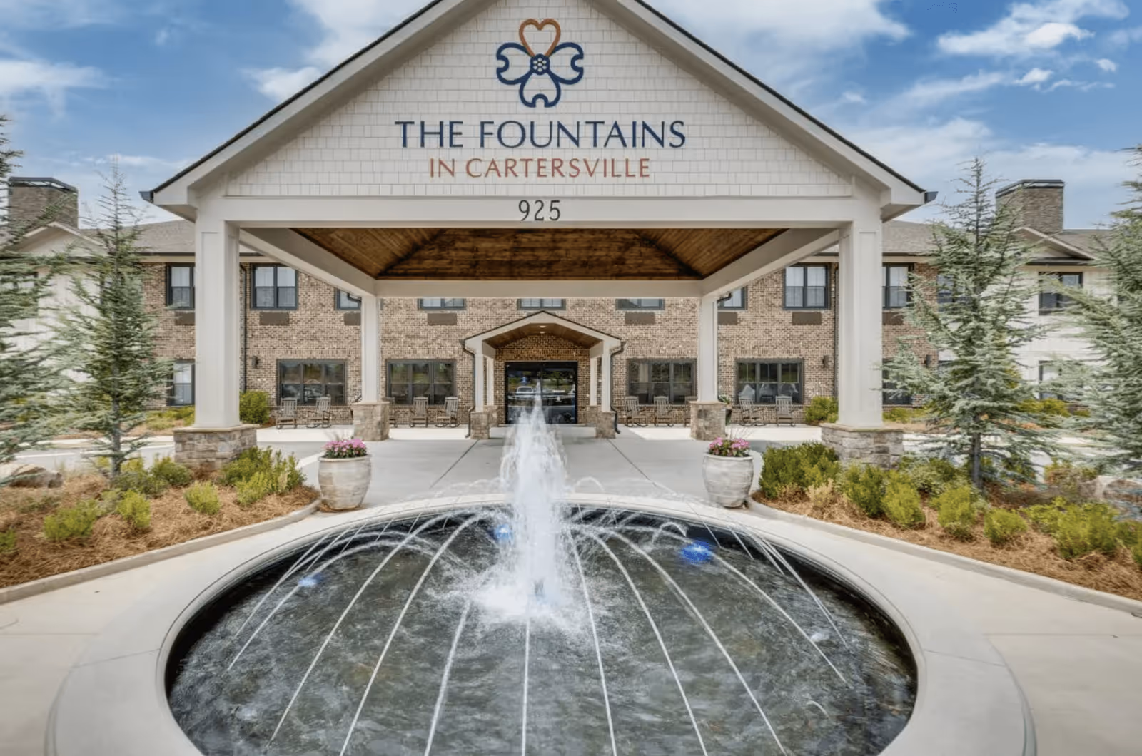 Front entrance of The Fountains in Cartersville assisted living facility featuring a covered driveway with a peaked roof, a water fountain in the foreground, landscaped greenery, and a brick building in the background under a partly cloudy sky.
