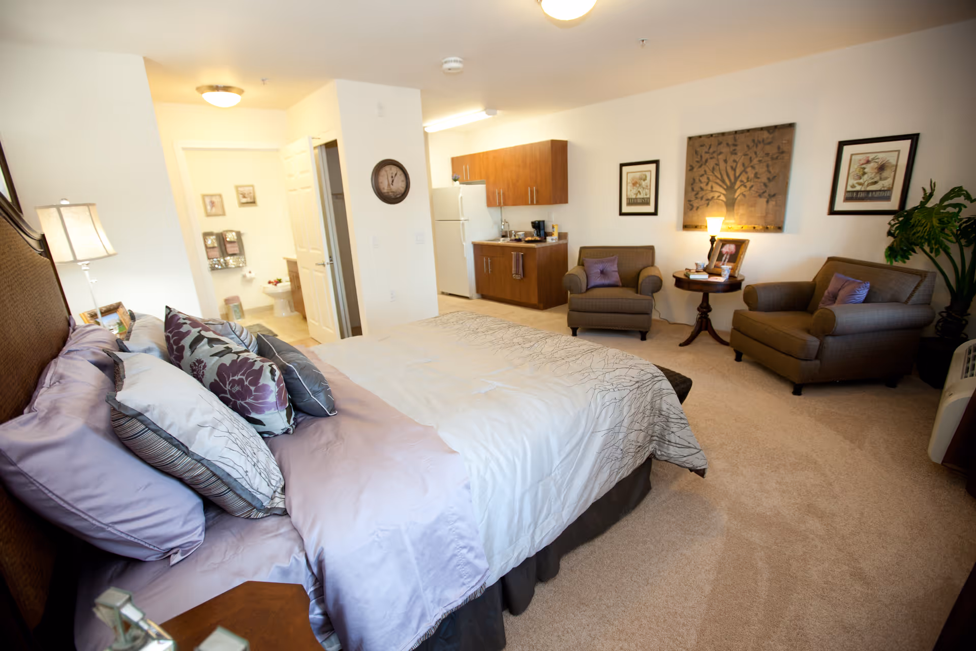 Bright furnished studio bedroom with a made bed, two armchairs and a small kitchenette in a senior living apartment.