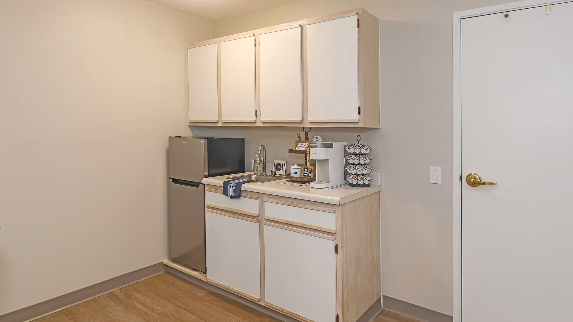 Small kitchenette area with white cabinets, a stainless steel mini refrigerator, a sink with a blue towel hanging over the edge, a coffee maker, and a rotating coffee pod holder. The walls are light beige, and there is a closed white door with a gold handle on the right side.