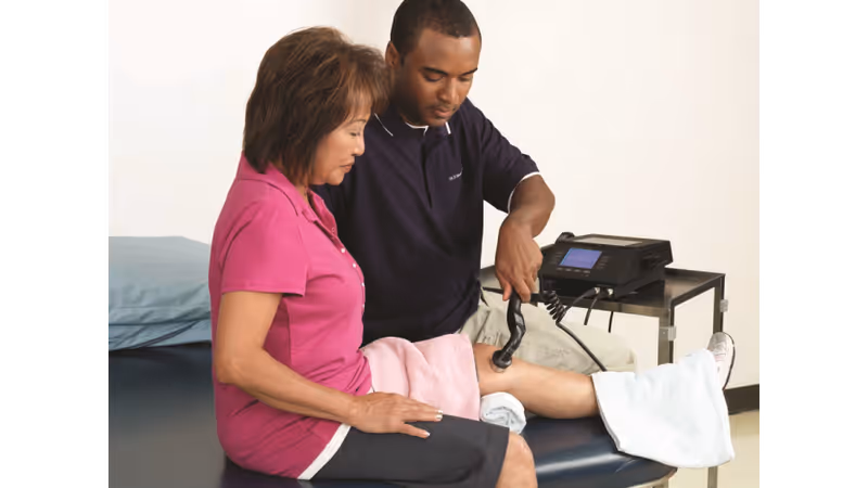 A healthcare professional is using a therapeutic ultrasound device on the knee of a seated woman in a clinical setting. The woman is wearing a pink shirt and black shorts, and her leg is covered with a towel. The healthcare professional is focused on the treatment, and medical equipment is visible on a small table nearby.