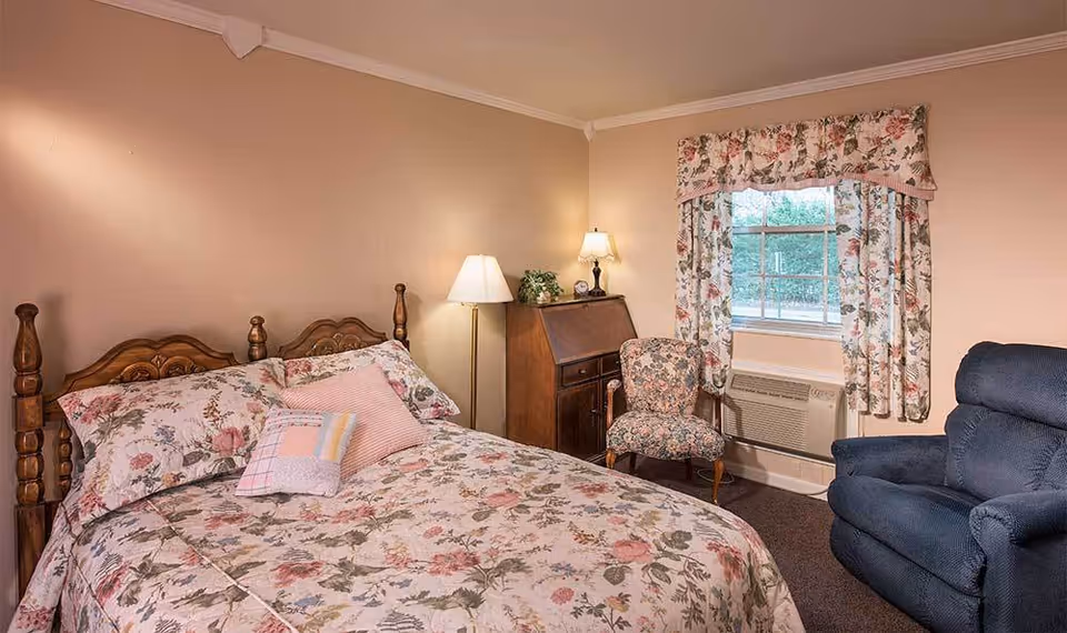 A cozy bedroom with a floral-patterned bedspread and pillows on a wooden bed frame. The room includes a floral upholstered chair, a blue recliner, a wooden desk with a lamp and plant, and a window with floral curtains. The walls are painted beige and the floor is carpeted.