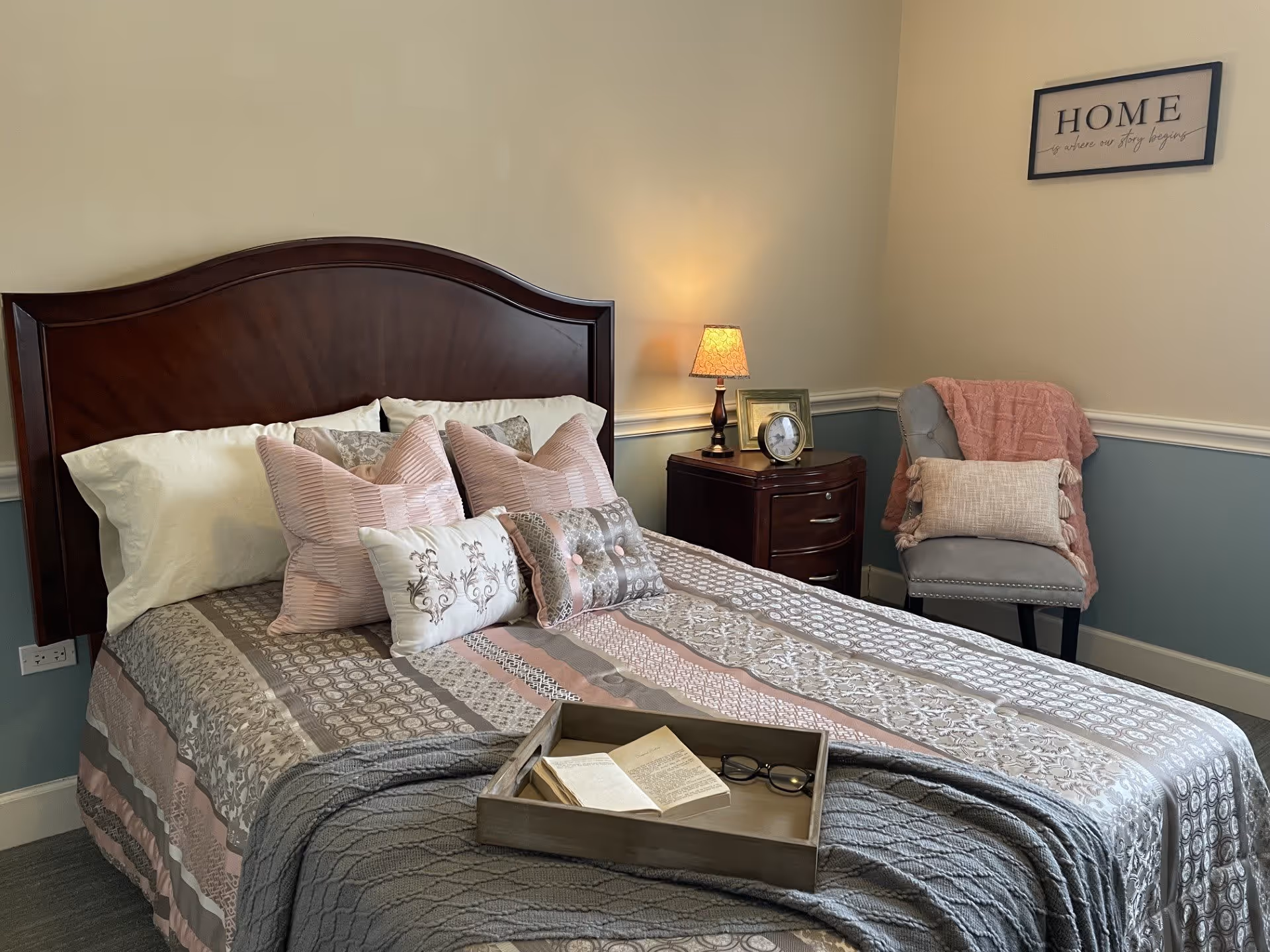 A neatly made bed with a wooden headboard, adorned with multiple decorative pillows in shades of pink, white, and gray. A wooden tray with an open book and eyeglasses rests on the bed. Next to the bed is a dark wooden nightstand with a lit lamp, a framed photo, and a clock. In the corner, there is a gray upholstered chair with a pink throw blanket and a beige pillow. A framed sign on the wall reads 'HOME is where our story begins.' The room has light-colored walls with a blue lower half and white trim.