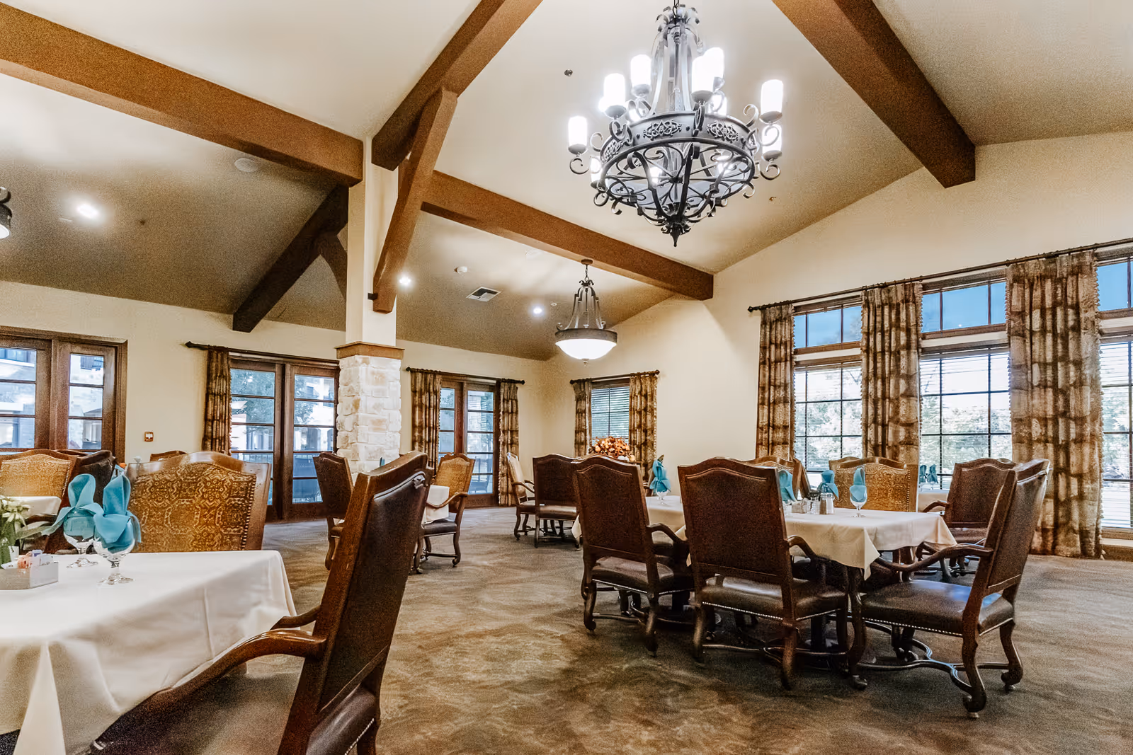 A spacious dining room with multiple tables covered in white tablecloths, each set with blue folded napkins and glassware. The room features large windows with patterned curtains, exposed wooden beams on the ceiling, and elegant chandeliers providing warm lighting. Comfortable wooden chairs surround the tables, and the overall ambiance is inviting and well-lit.