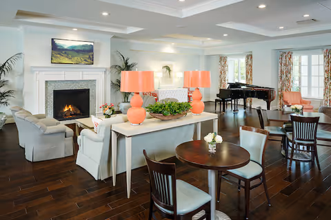 A spacious and well-lit living room area with hardwood floors, featuring a fireplace with a TV mounted above it, a grand piano near large windows with floral curtains, several seating arrangements including sofas and armchairs, round wooden tables with chairs, and a console table with two orange lamps and a plant centerpiece.