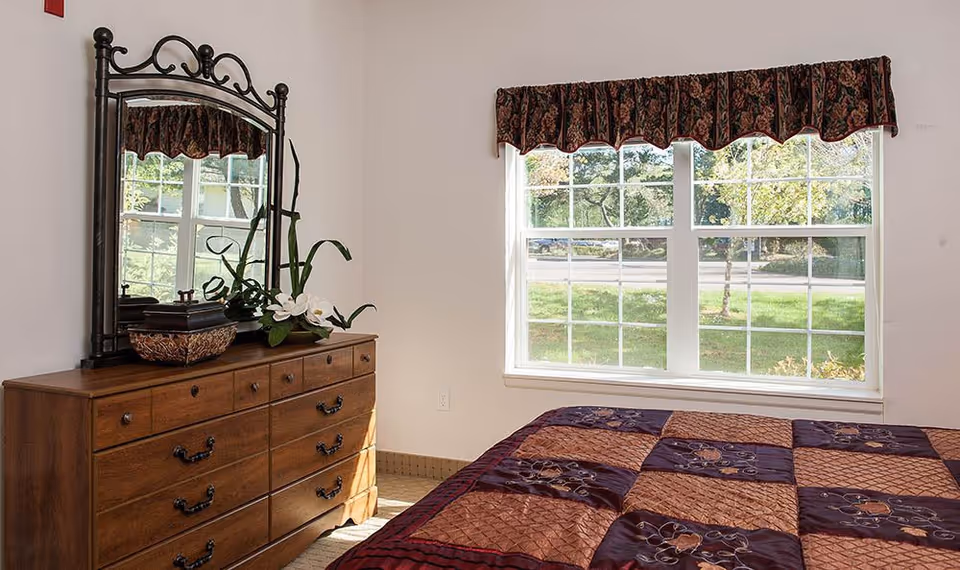 Sunlit bedroom with a wooden dresser topped by an ornate mirror and plants, a patterned quilted bed, and a large window overlooking a green lawn.