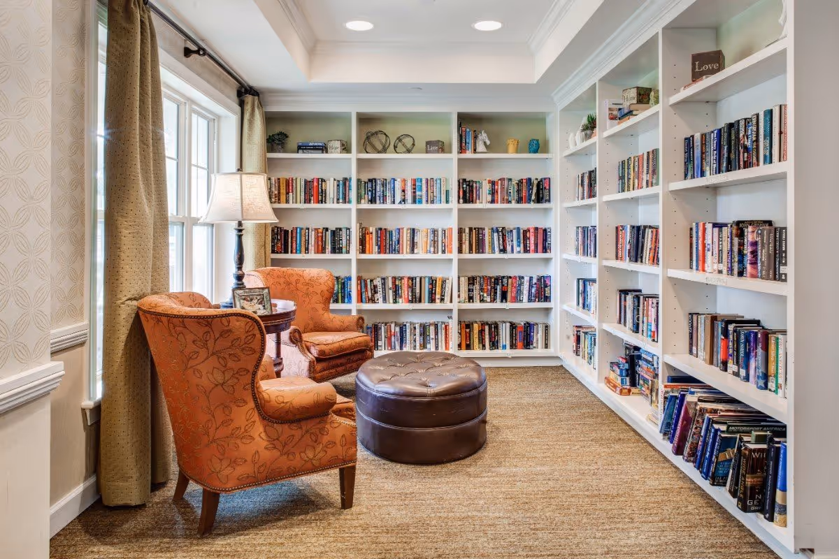 A cozy reading nook in a senior living facility featuring two orange patterned armchairs, a round brown leather ottoman, a small round wooden table with a lamp and picture frame, and white built-in bookshelves filled with books along two walls. Natural light comes in through a window with beige curtains.