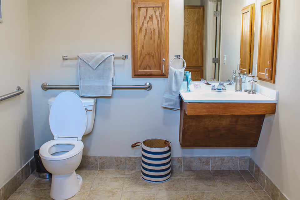 A clean bathroom with a white toilet, a towel hanging on a metal grab bar behind the toilet, a wooden cabinet mounted on the wall, a white sink with a wooden base, a mirror above the sink, and a striped basket on the tiled floor.
