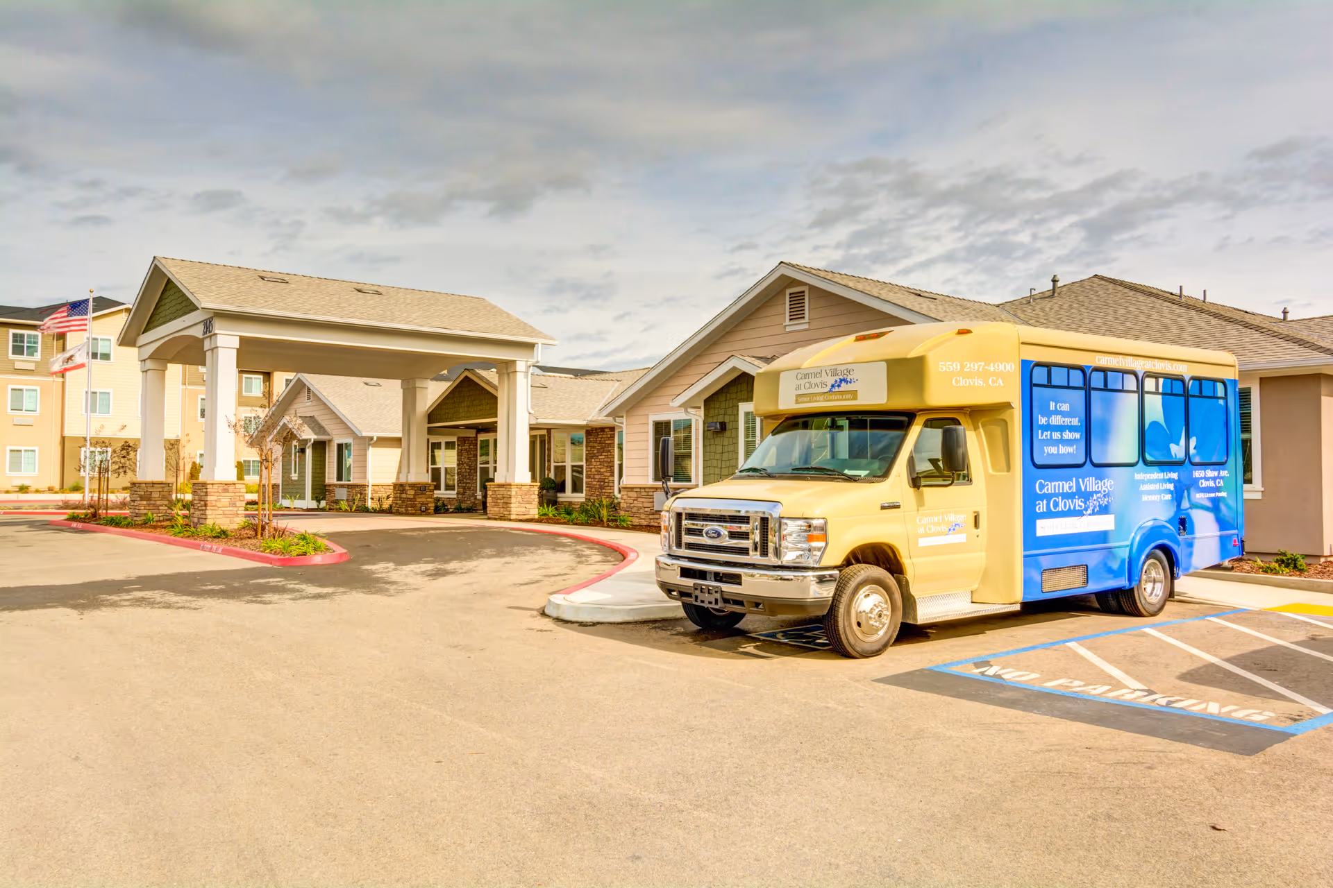 Exterior view of Carmel Village Memory Care facility showing the main entrance with a covered drop-off area and a yellow and blue shuttle bus parked in a handicapped parking space. The building has beige and green siding with stone accents and an American flag is visible in the background.