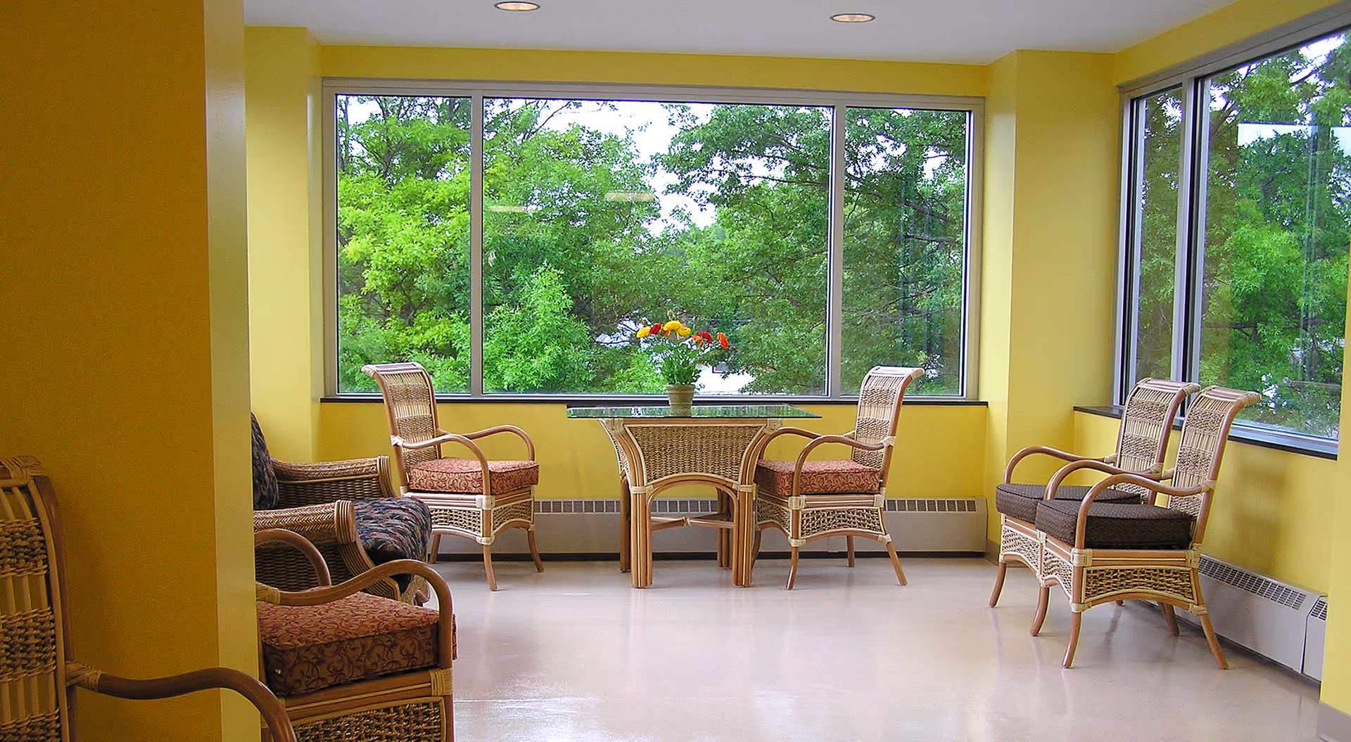 Sunlit sitting area with wicker chairs and a small table topped with flowers in front of large windows overlooking trees.