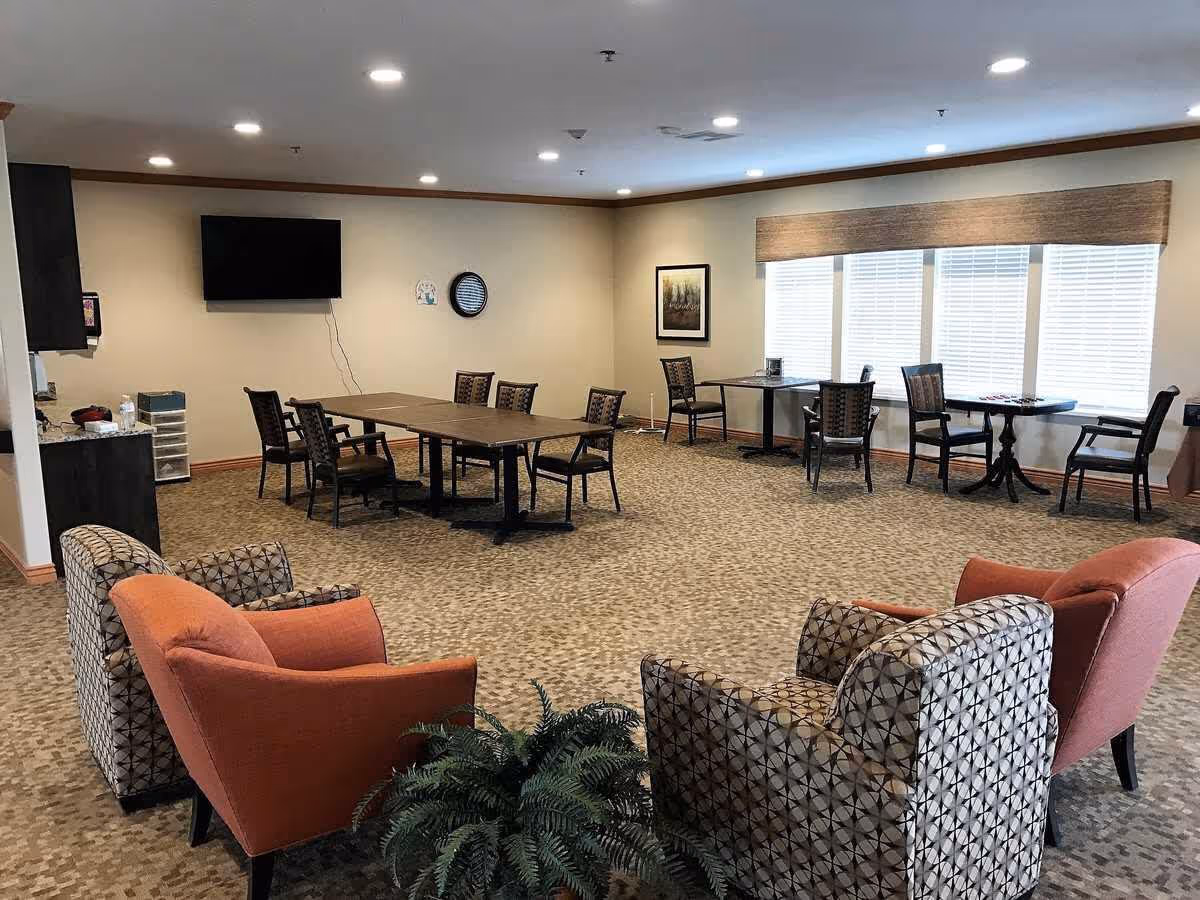 A spacious assisted-living common room with multiple chairs, tables, a wall-mounted TV and large windows.