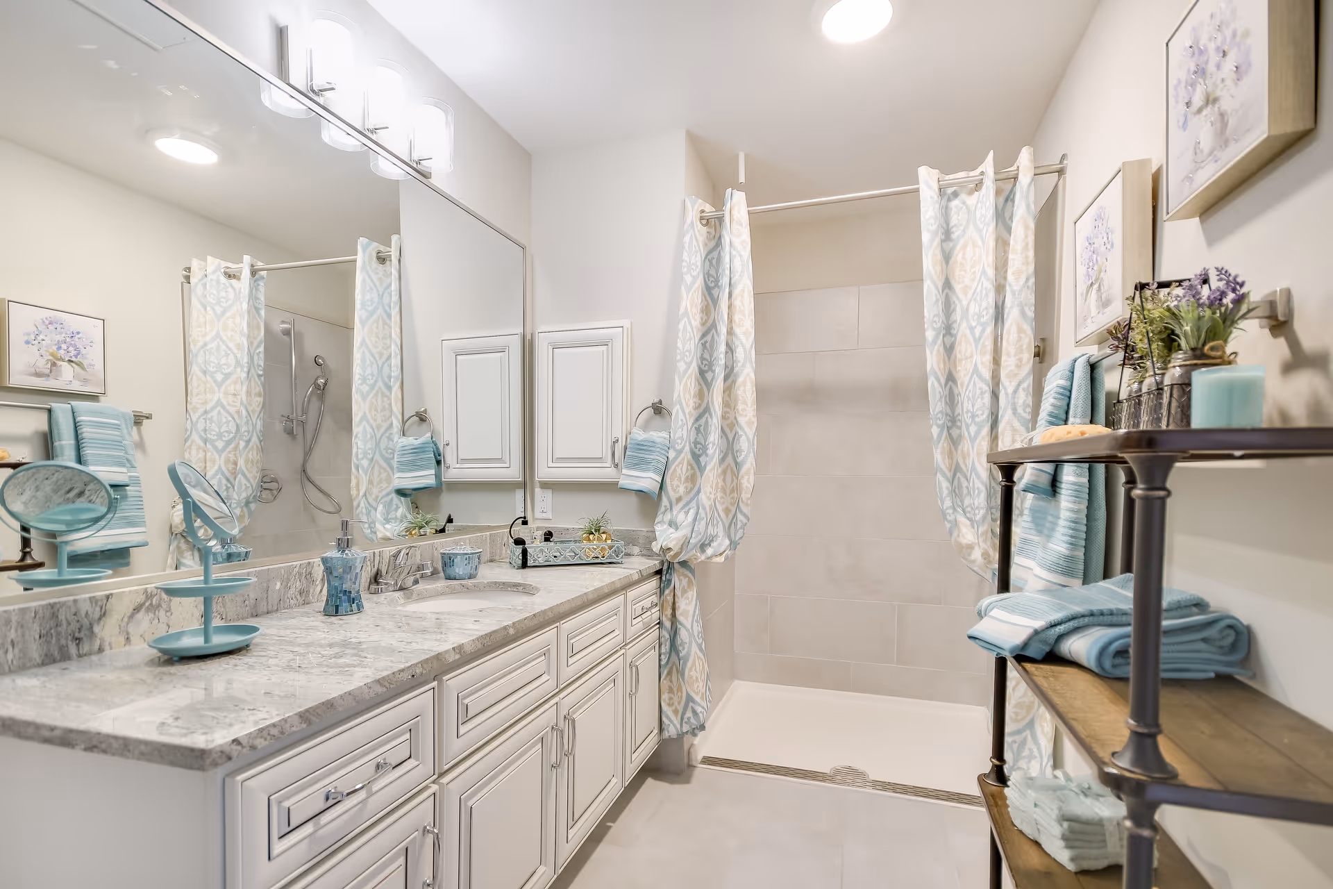 A bright and clean bathroom featuring a large marble countertop with a sink, a wide mirror above the counter, and a walk-in shower with a patterned shower curtain. The bathroom is decorated with blue and white towels, small plants, and framed floral artwork on the walls. There is a wooden shelving unit holding additional towels and decorative items.