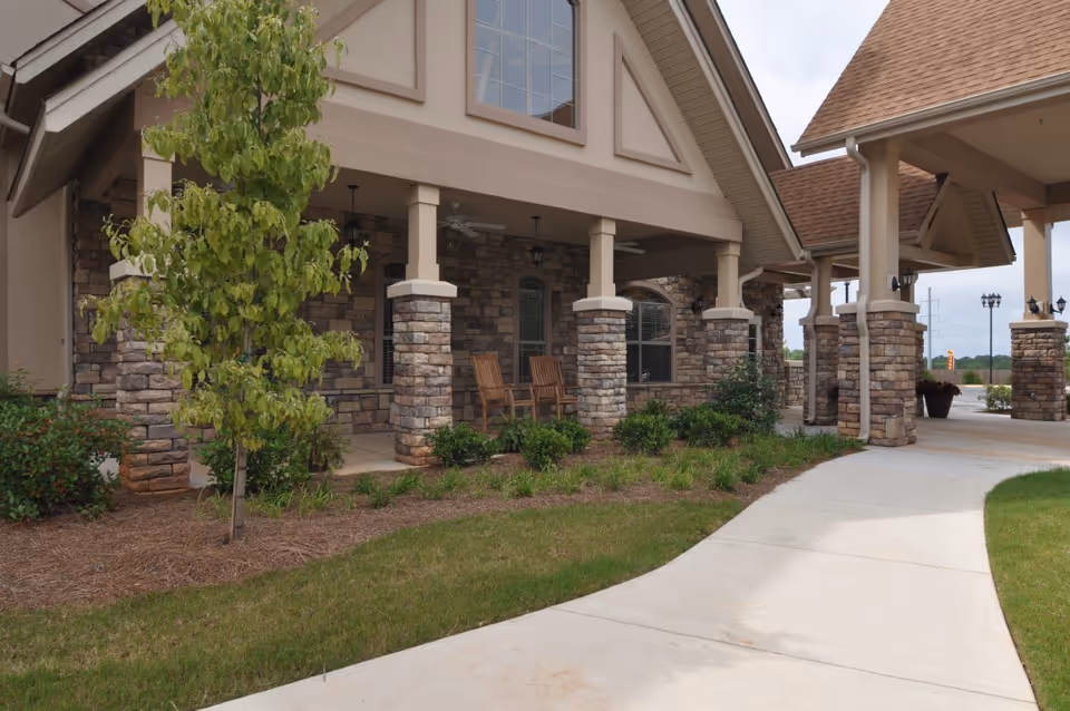 Front entrance of a senior living building with stone columns, a covered porch with rocking chairs, landscaping and a curved sidewalk.