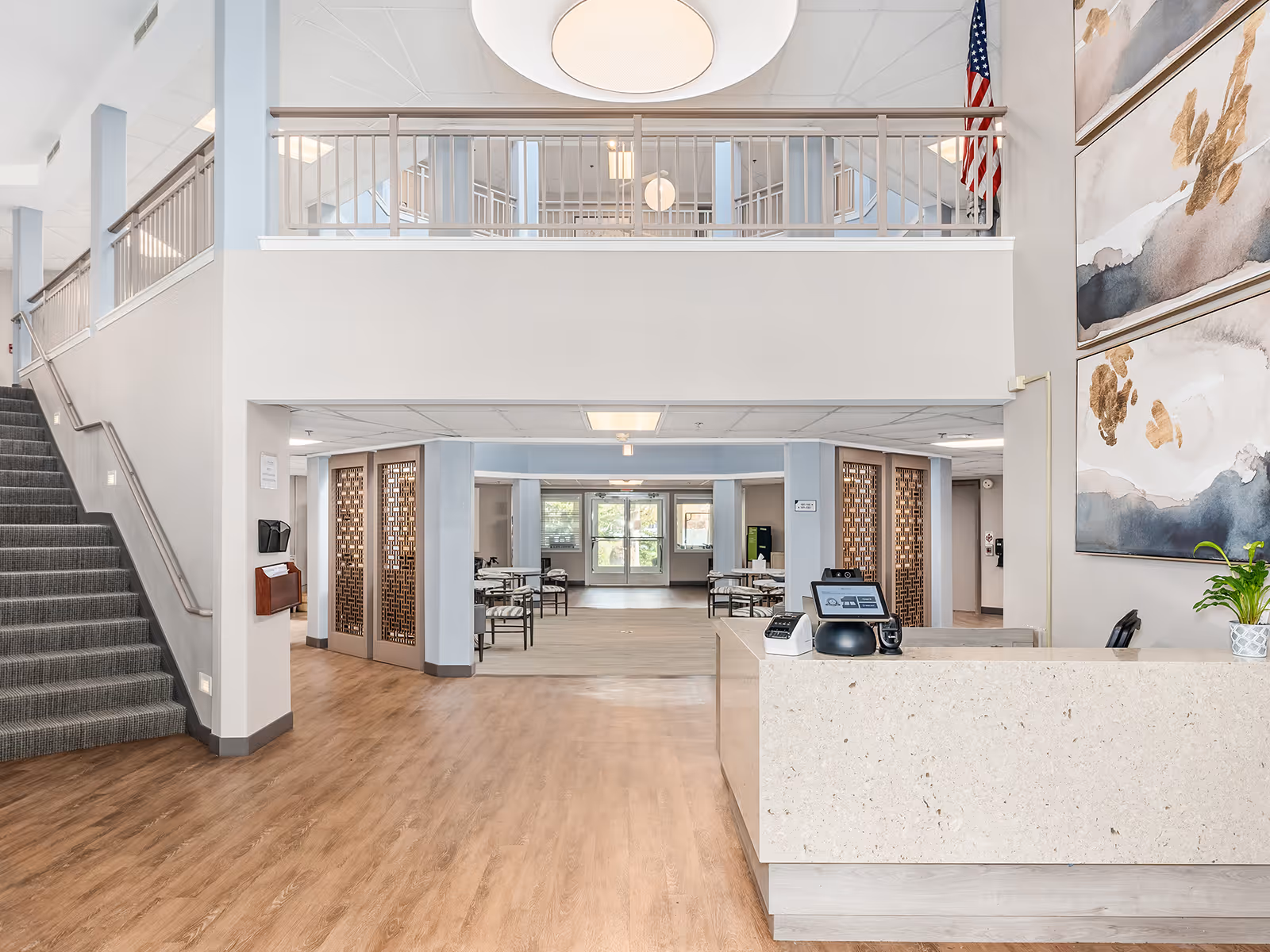 Bright and spacious senior living facility lobby with a reception desk on the right, a staircase on the left, and a seating area with tables and chairs in the background. The area features wood flooring, light-colored walls, large abstract paintings, and an American flag hanging near the ceiling.