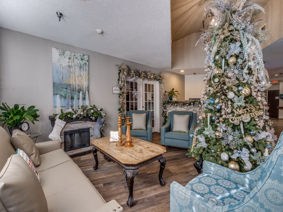 A cozy living room decorated for Christmas with a large Christmas tree adorned with ornaments and a 'Merry Christmas' sign. The room features a beige sofa, two blue armchairs, a wooden coffee table with candle holders, a fireplace with stockings and garland, and a painting of trees on the wall.