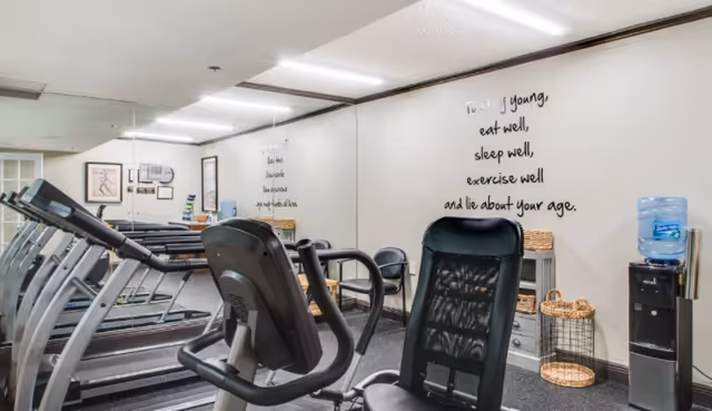 Indoor fitness room with several treadmills lined up against a mirrored wall. A stationary exercise bike is in the foreground. On the right wall, there is a motivational quote that reads: 'To stay young, eat well, sleep well, exercise well and lie about your age.' A water cooler and a basket are also visible near the wall.