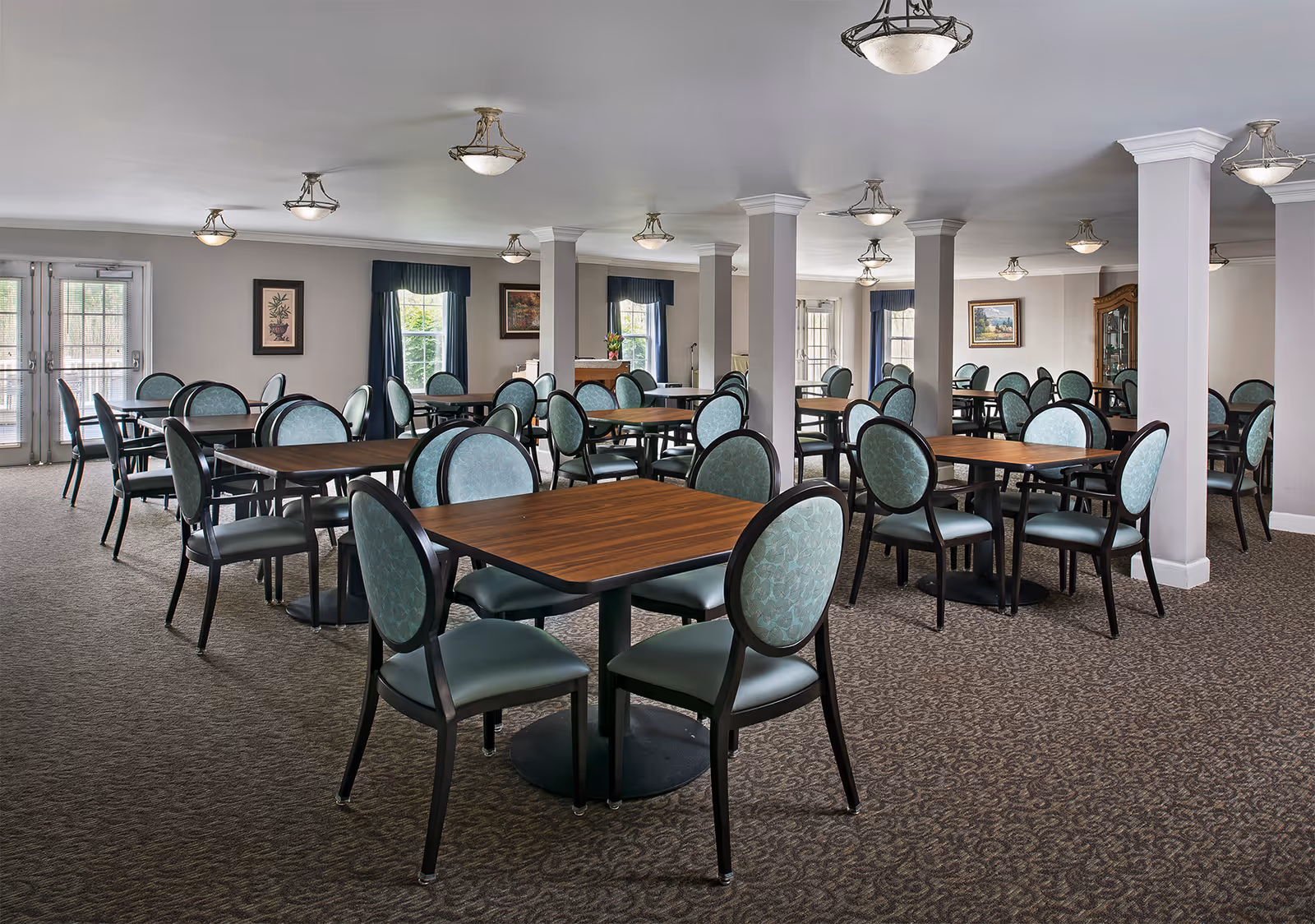 A spacious dining room with multiple wooden tables and green cushioned chairs arranged neatly. The room has beige walls, carpeted floors, several windows with dark green curtains, and ceiling light fixtures. There are framed pictures on the walls and white pillars throughout the room.