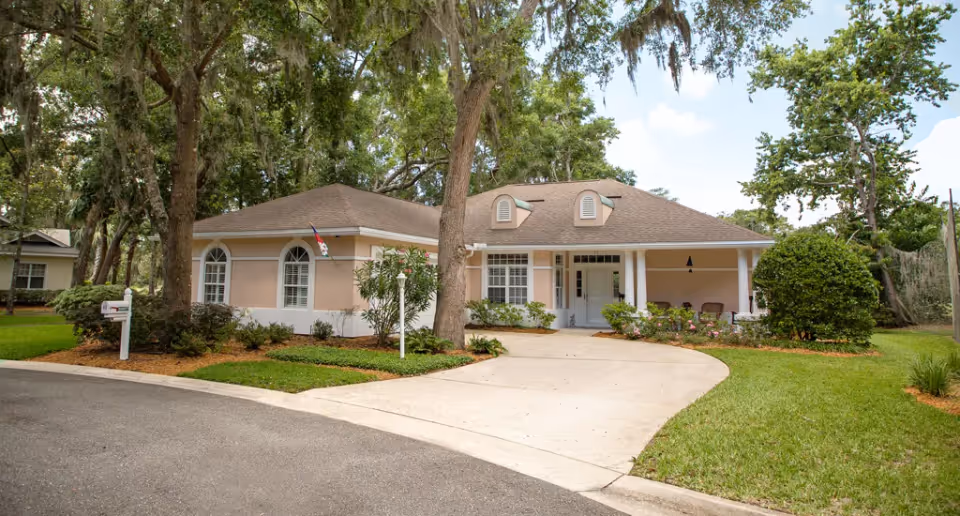 Single-story beige house with a curved driveway, front porch, and surrounding trees and lawn.