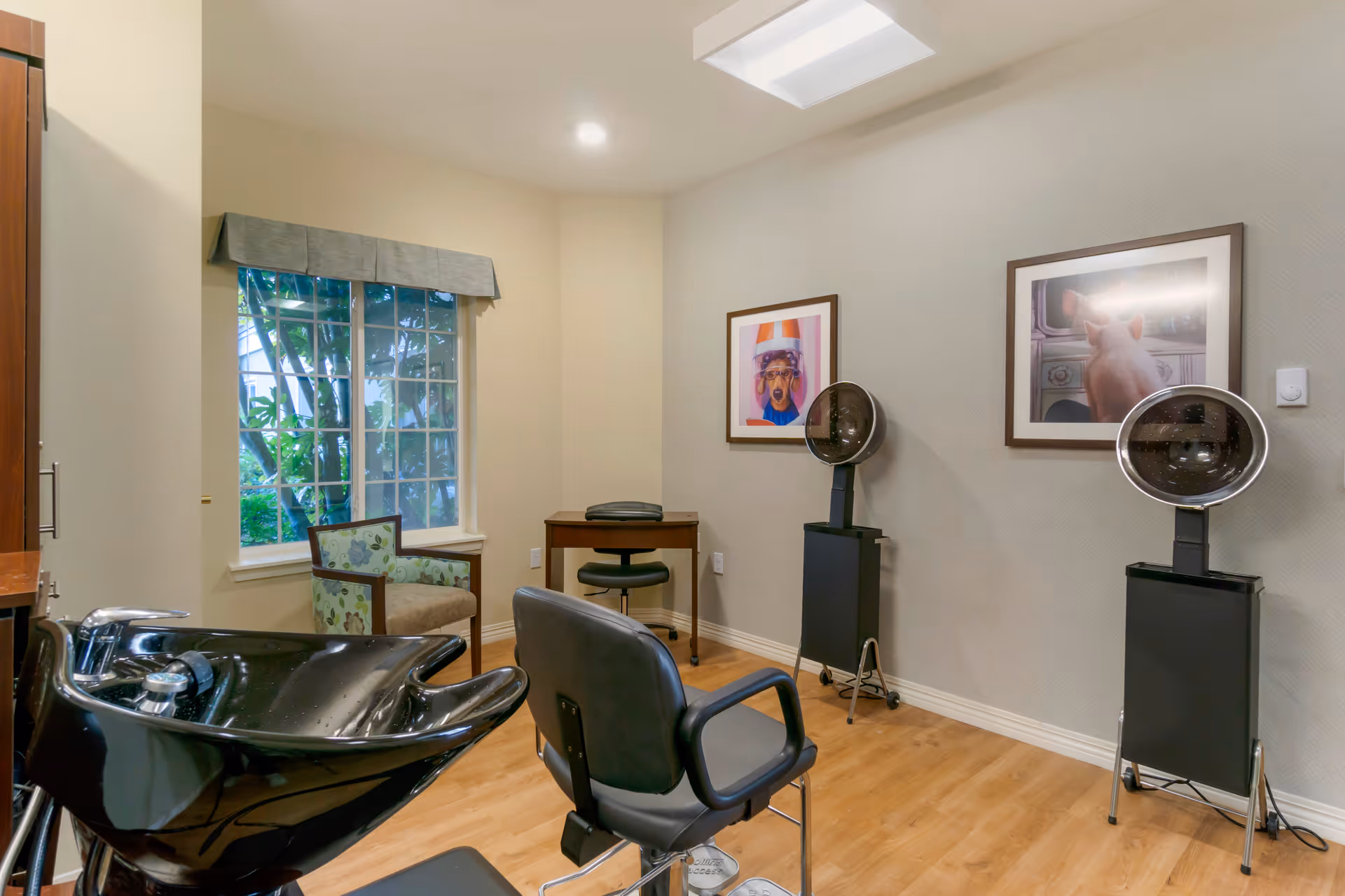 Interior view of a hair salon area in a senior living facility with a black salon chair and wash basin in the foreground, two hair drying stations against the wall, a small desk with a chair, a cushioned armchair near a window with a valance, and two framed pictures on the wall.
