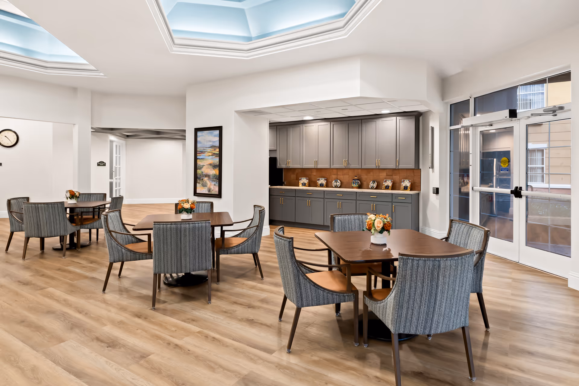 A bright and spacious common dining area with several wooden tables and cushioned chairs arranged neatly. Each table has a small floral centerpiece. The room features light wood flooring, a large skylight ceiling, and a wall with gray cabinets and decorative jars. There is a glass door leading outside on the right side.