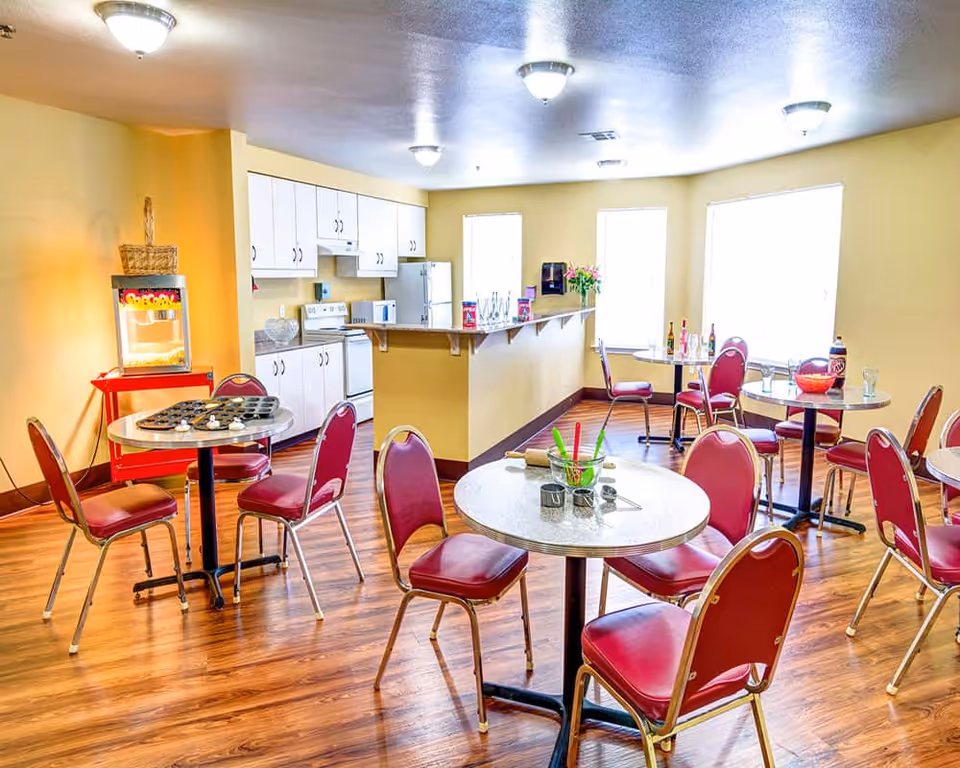 A bright dining area with multiple round tables and red chairs on a wooden floor. The room has yellow walls and several windows letting in natural light. In the background, there is a kitchen area with white cabinets, a stove, microwave, and refrigerator. A popcorn machine is visible on a red cart near one of the tables.