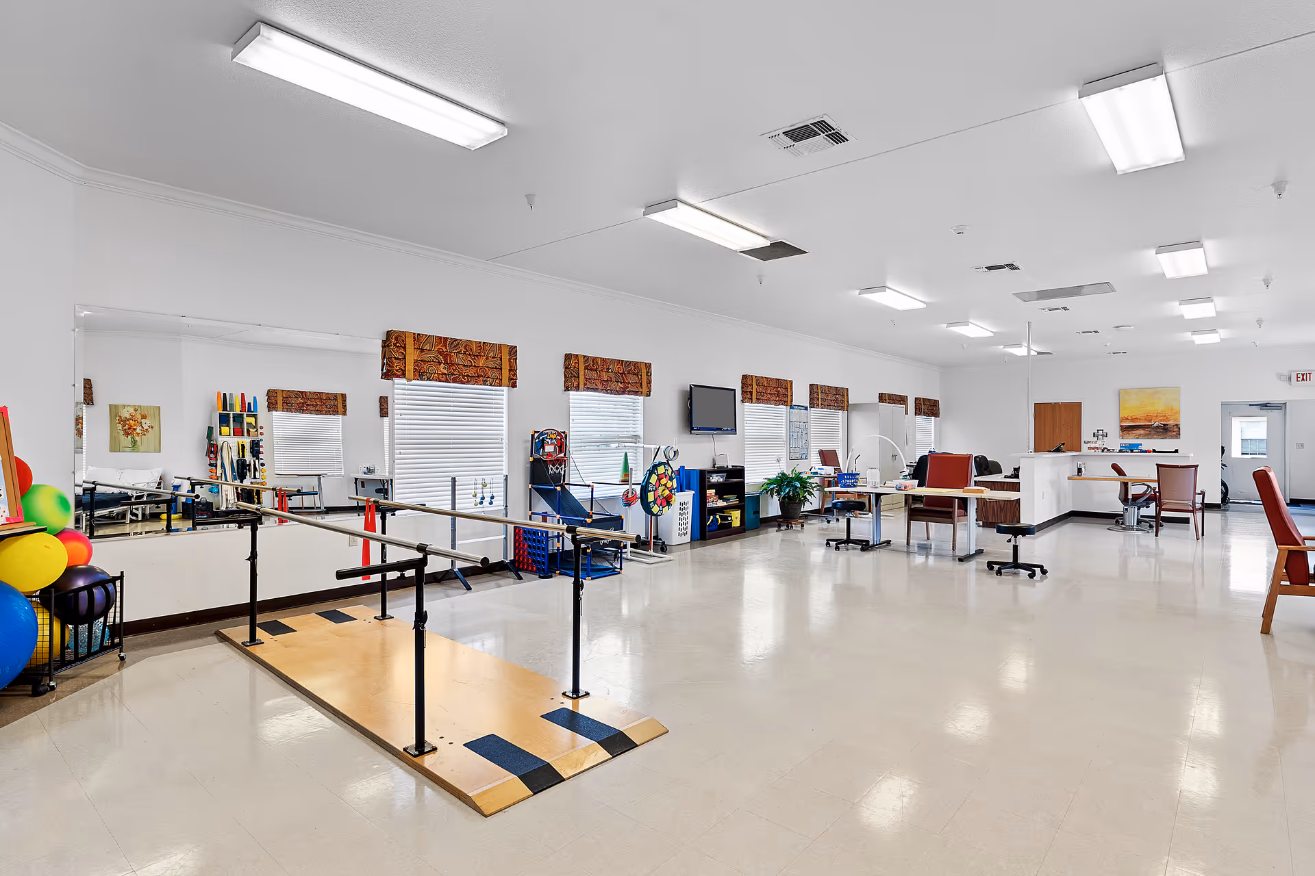 A spacious rehabilitation room with parallel bars for walking exercises, colorful therapy balls, a large mirror on the wall, several windows with patterned valances, chairs, tables, and therapy equipment. The room is well-lit with fluorescent ceiling lights and has a clean, polished floor.