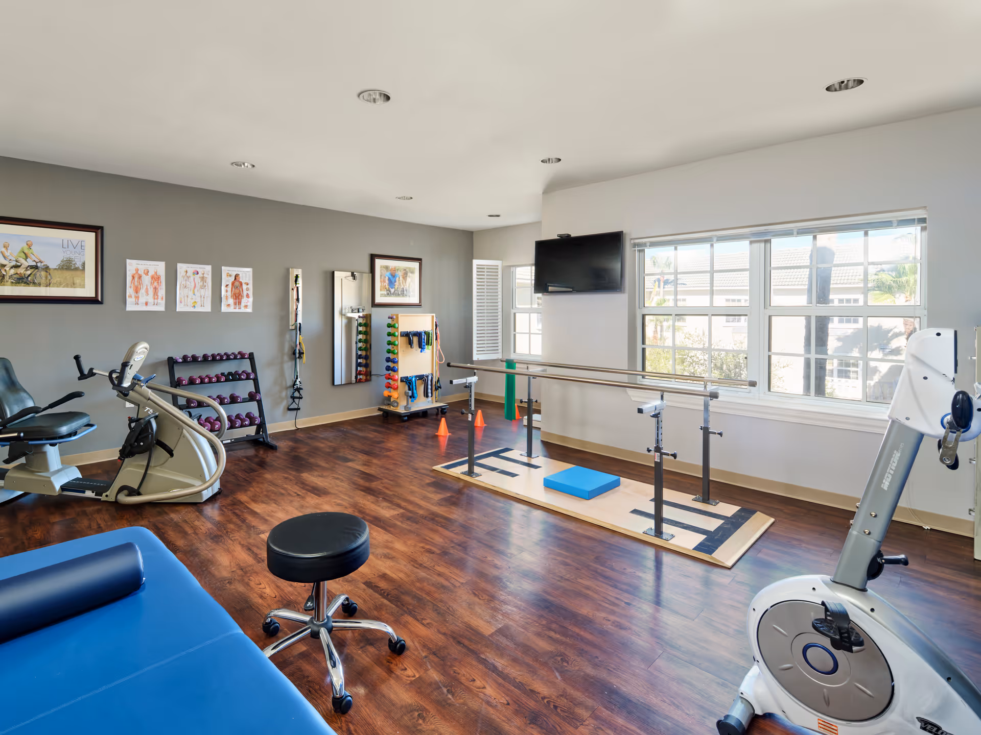 Bright rehabilitation gym room with exercise bikes, free weights, parallel bars, and a TV by a large window.