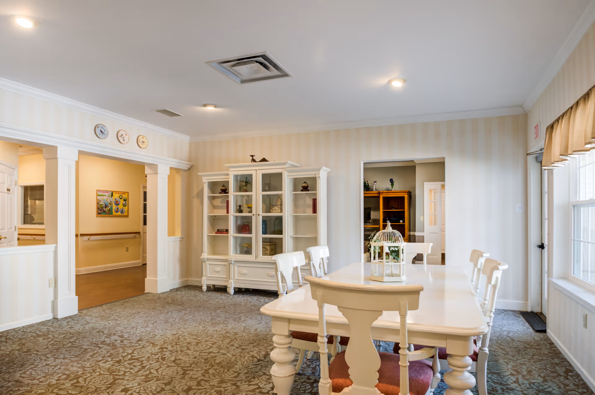 Bright communal dining room with a white table and chairs, a glass-front cabinet, and windows letting in natural light.