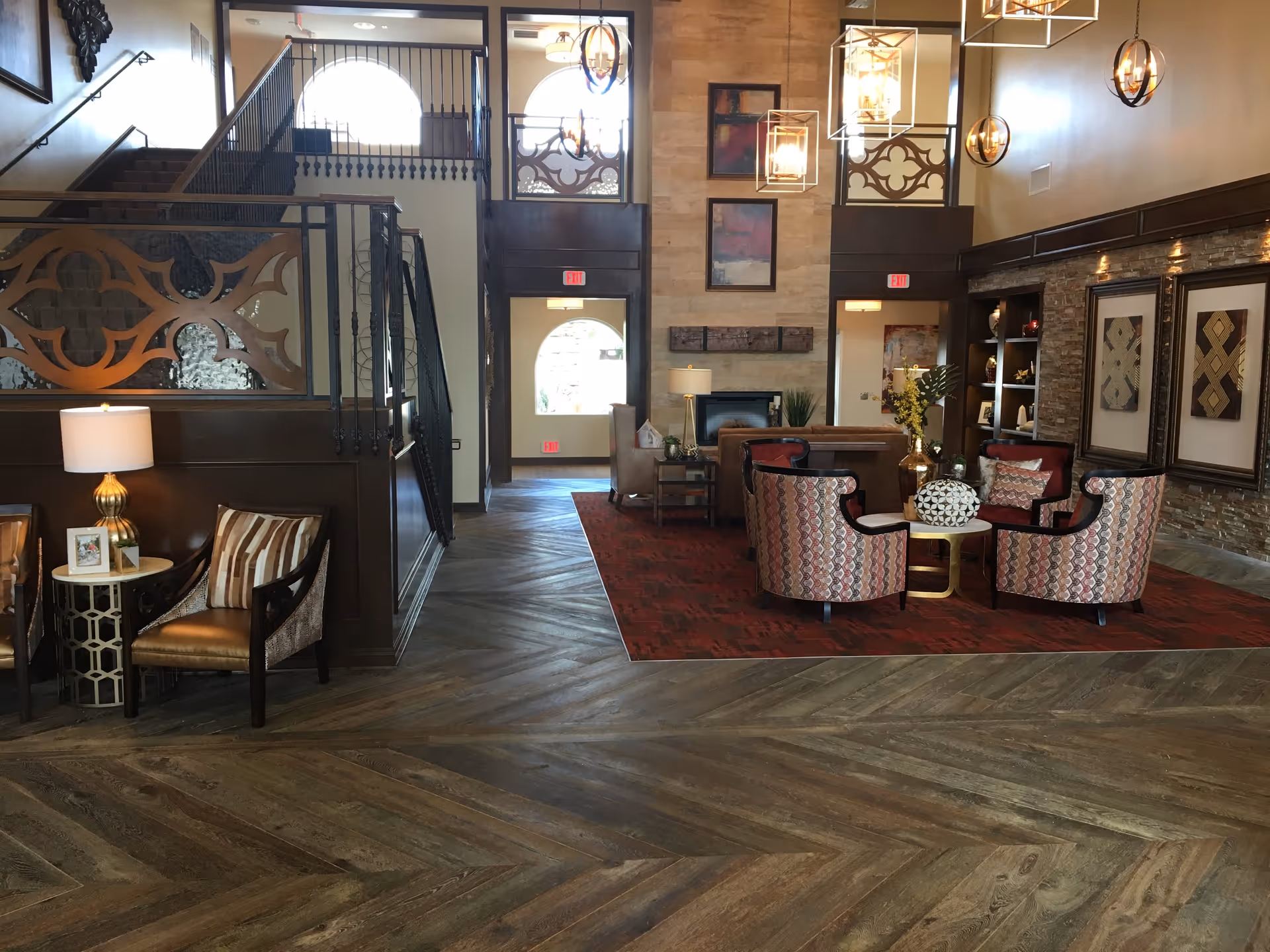 A spacious and elegantly decorated living room area with patterned armchairs arranged around a small round table on a red rug. The room features wooden flooring, a stone accent wall with framed artwork, and modern hanging light fixtures. There is a staircase with decorative railing on the left and large windows allowing natural light to enter.