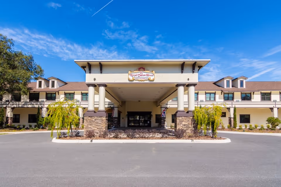 Front exterior view of Royal Dalton House, a two-story building with a covered entrance supported by columns, surrounded by landscaping and a clear blue sky above.