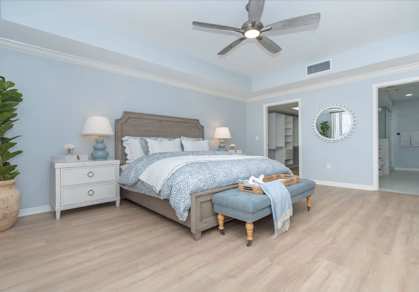 Bright master bedroom with a large bed, nightstands, bench at foot, ceiling fan, and open doorways to a bathroom and closet.