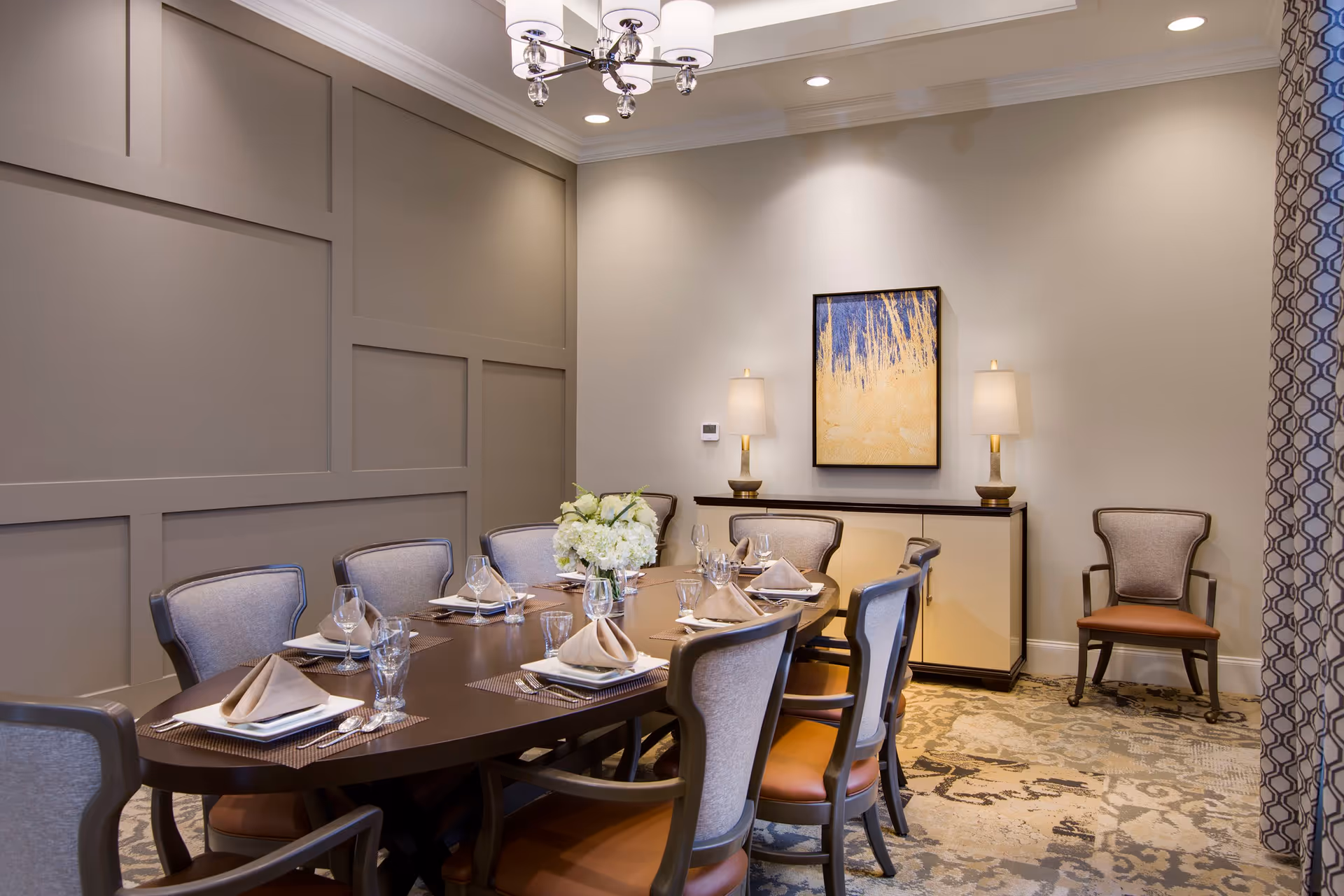 A formal dining room with a long dark wooden table set for eight people. Each place setting includes a folded beige napkin, a plate, a glass, and silverware. The chairs have cushioned seats and backs in neutral tones. A floral centerpiece decorates the middle of the table. The room features a patterned carpet, a sideboard with two lamps, a framed abstract painting on the wall, and a modern chandelier hanging from the ceiling. A patterned curtain is partially visible on the right side.
