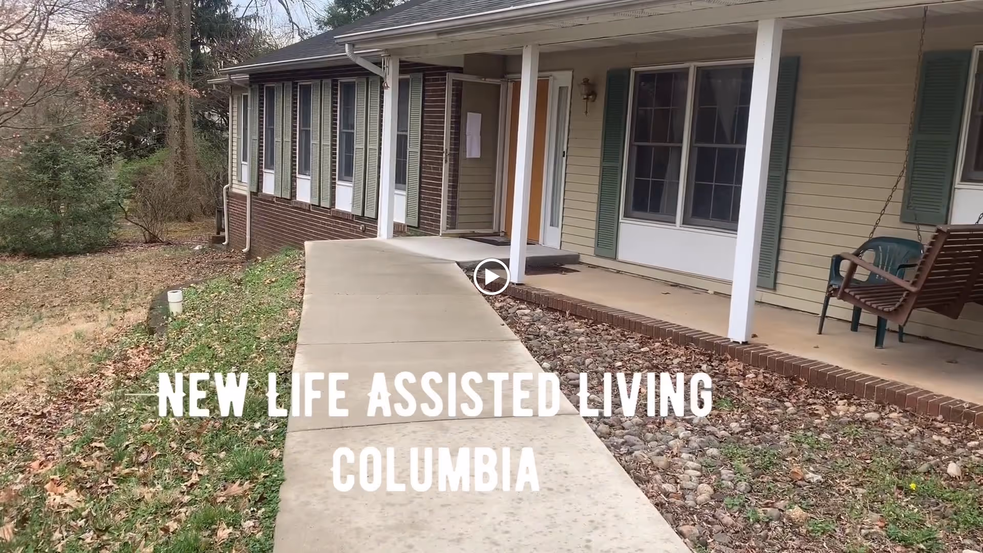 Exterior view of a single-story assisted living facility with a concrete walkway leading to the entrance. The building has beige siding, green shutters, and a covered porch with a wooden swing and a plastic chair. Surrounding the walkway are grass and some trees.