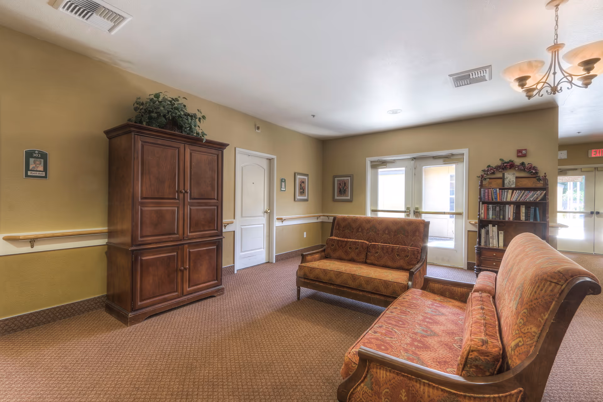 A cozy common area in a senior living facility featuring two patterned upholstered sofas facing each other, a wooden cabinet with a plant on top, a bookshelf filled with books, beige walls with framed artwork, and double glass doors leading outside. The room has carpeted flooring and ceiling lights.