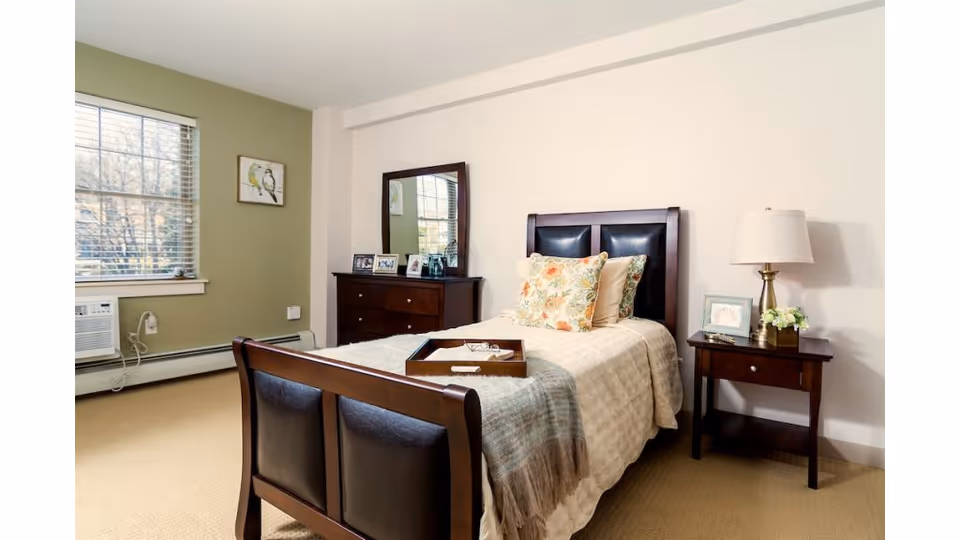 A cozy senior living bedroom with a single bed featuring floral pillows and a beige quilt. There is a wooden nightstand with a lamp and a small flower arrangement on the right side of the bed. A wooden dresser with a mirror and framed photos is against the wall. A window with blinds and an air conditioning unit is on the left wall, and a small framed bird picture hangs next to the window.