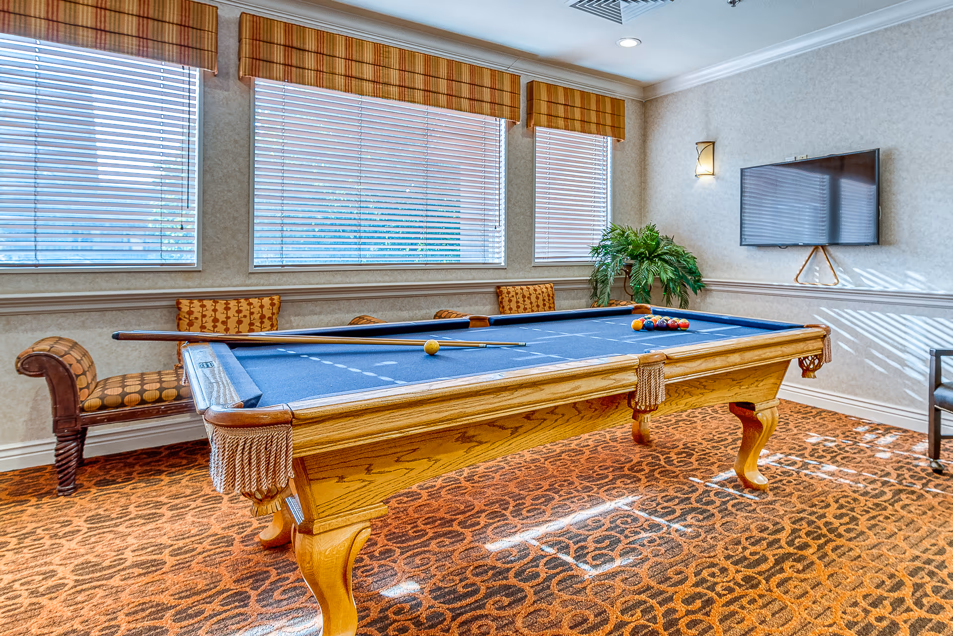 A well-lit game room with a wooden pool table featuring a blue felt surface. The pool table has a cue stick resting on it and a set of billiard balls arranged for a game. The room has large windows with blinds and striped valances, patterned carpet, a wall-mounted flat-screen TV, a green potted plant, and cushioned chairs along the wall.