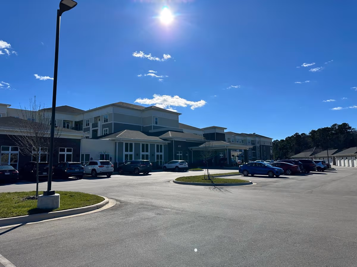 Exterior front of a multi-story senior living facility with a parking lot and cars under a bright sunny sky.