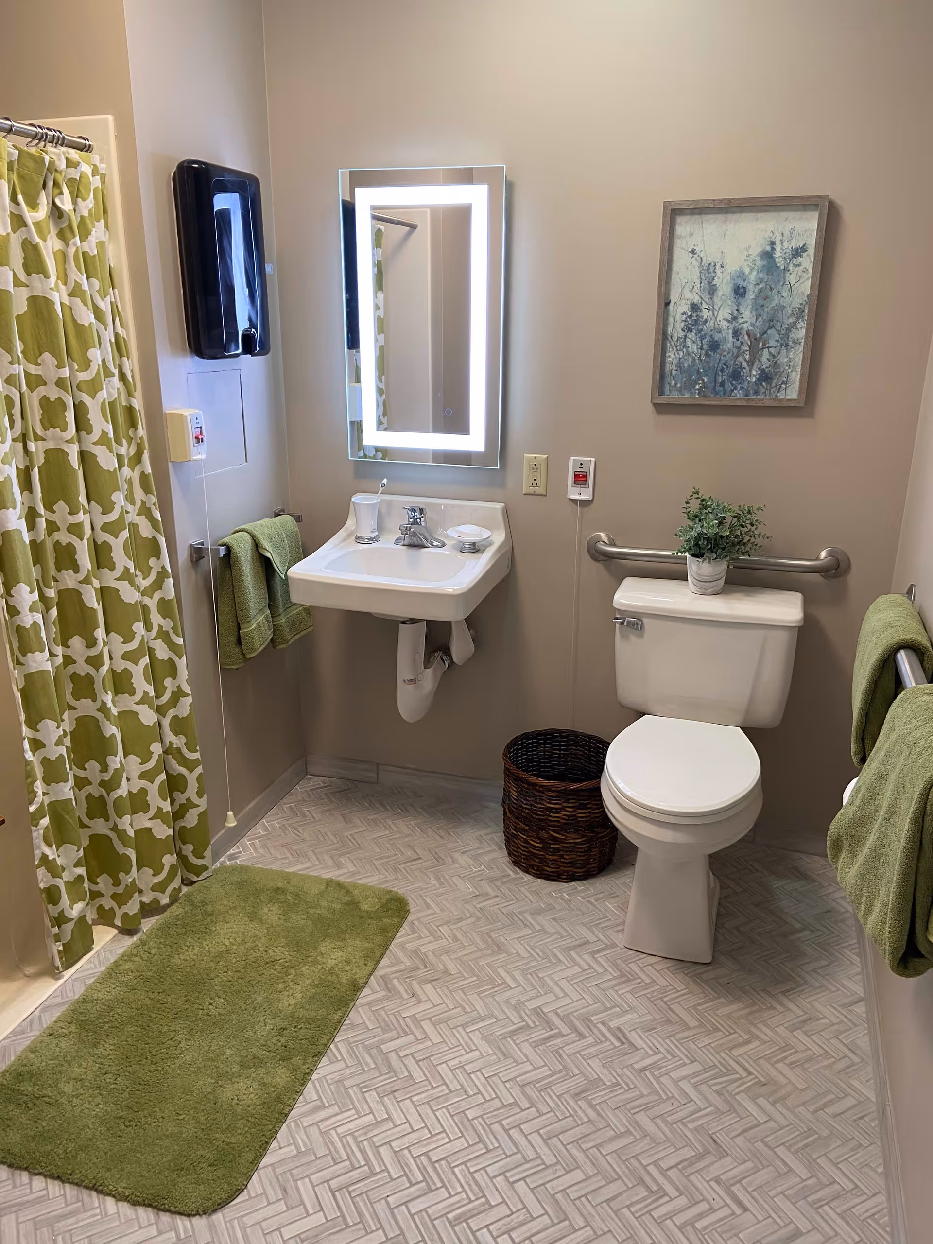 A clean bathroom with a white toilet, a small white sink with a modern rectangular mirror above it, a green patterned shower curtain, green bath mat, and green towels. There is a small plant on the toilet tank and a framed picture on the wall. The floor has a light herringbone pattern.