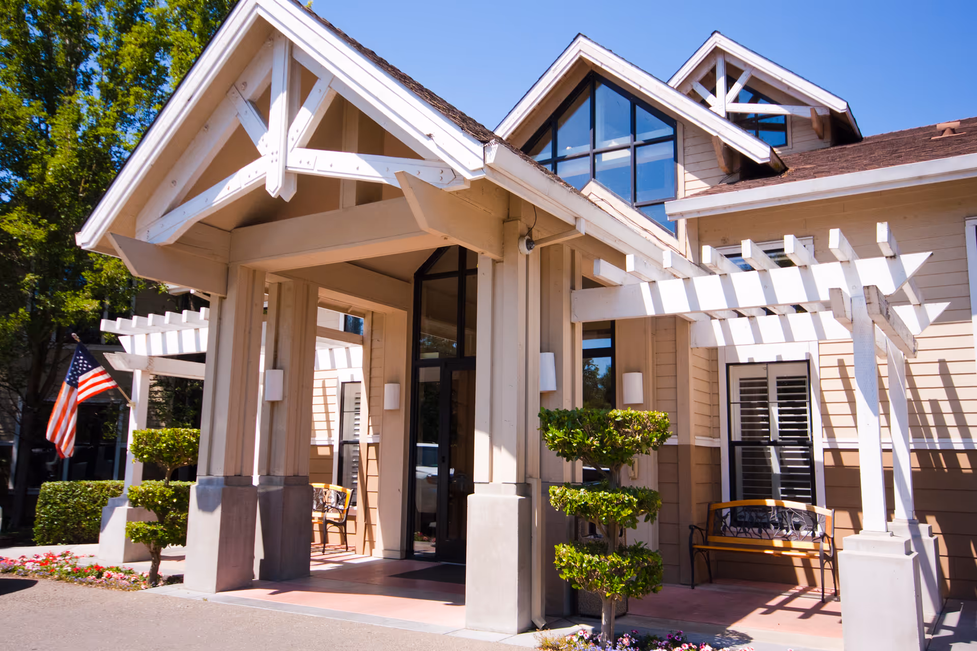 Covered entrance of a beige senior living building with a white pergola, manicured shrubs, an American flag, and a bench.