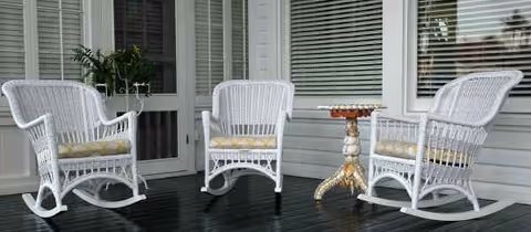 Three white wicker chairs and a small ornate table on a covered porch.