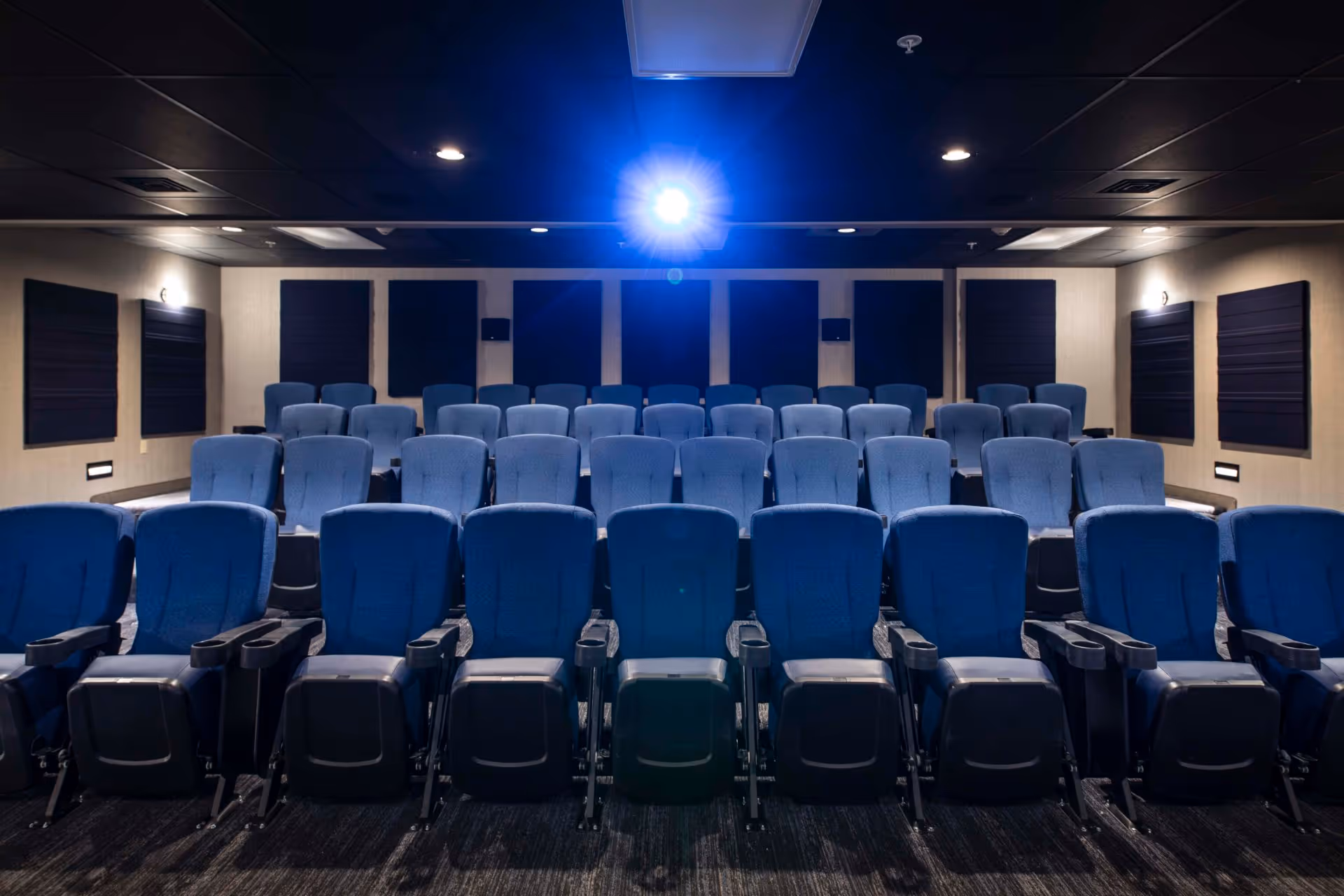 Interior view of a small theater or screening room with rows of blue cushioned seats facing a projector that is turned on, mounted on the ceiling. The walls have soundproof panels and the room is dimly lit.