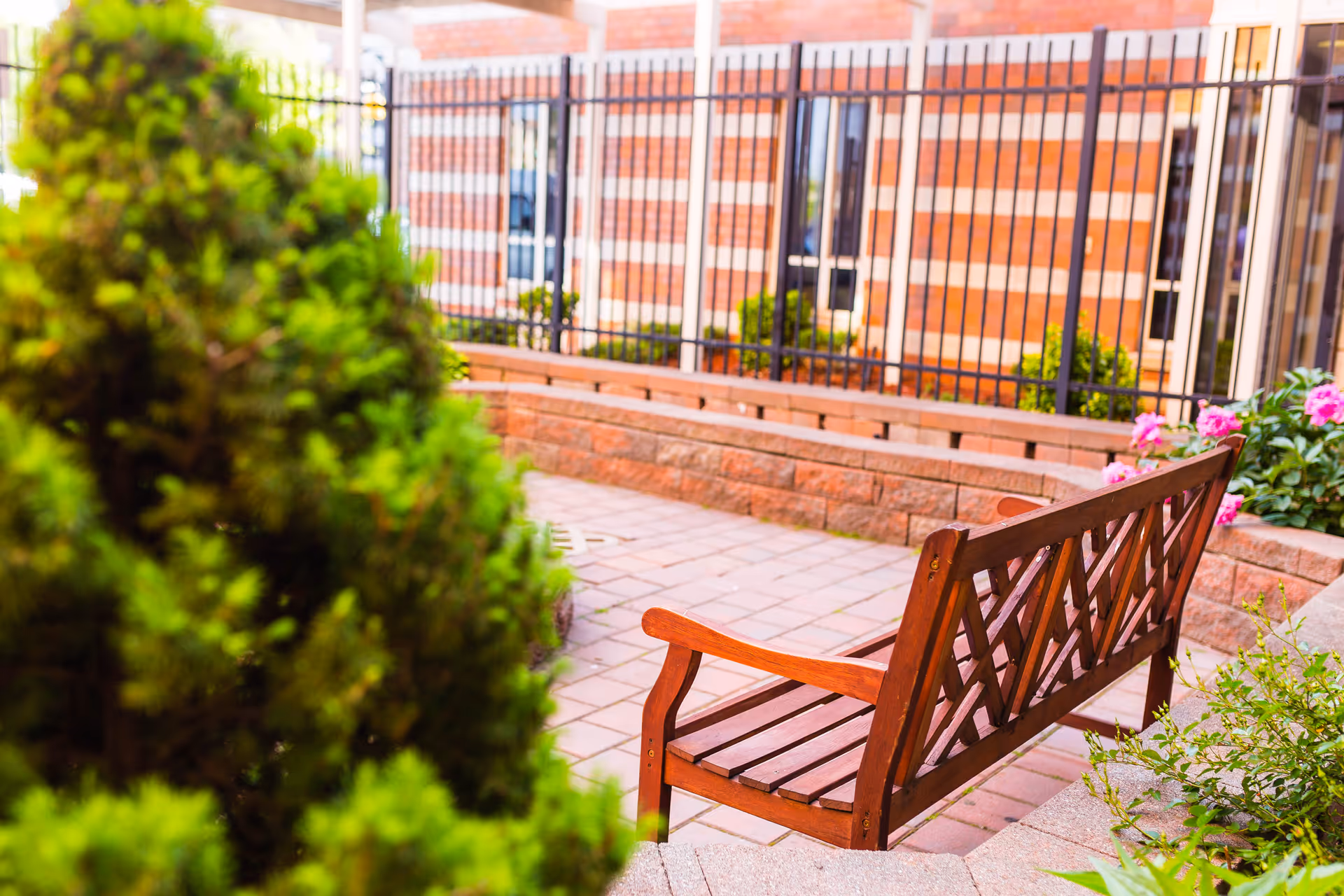 A wooden bench with a lattice backrest is placed on a paved patio area surrounded by green bushes and pink flowers. Behind the bench, there is a brick wall with a black metal fence and windows of a building.