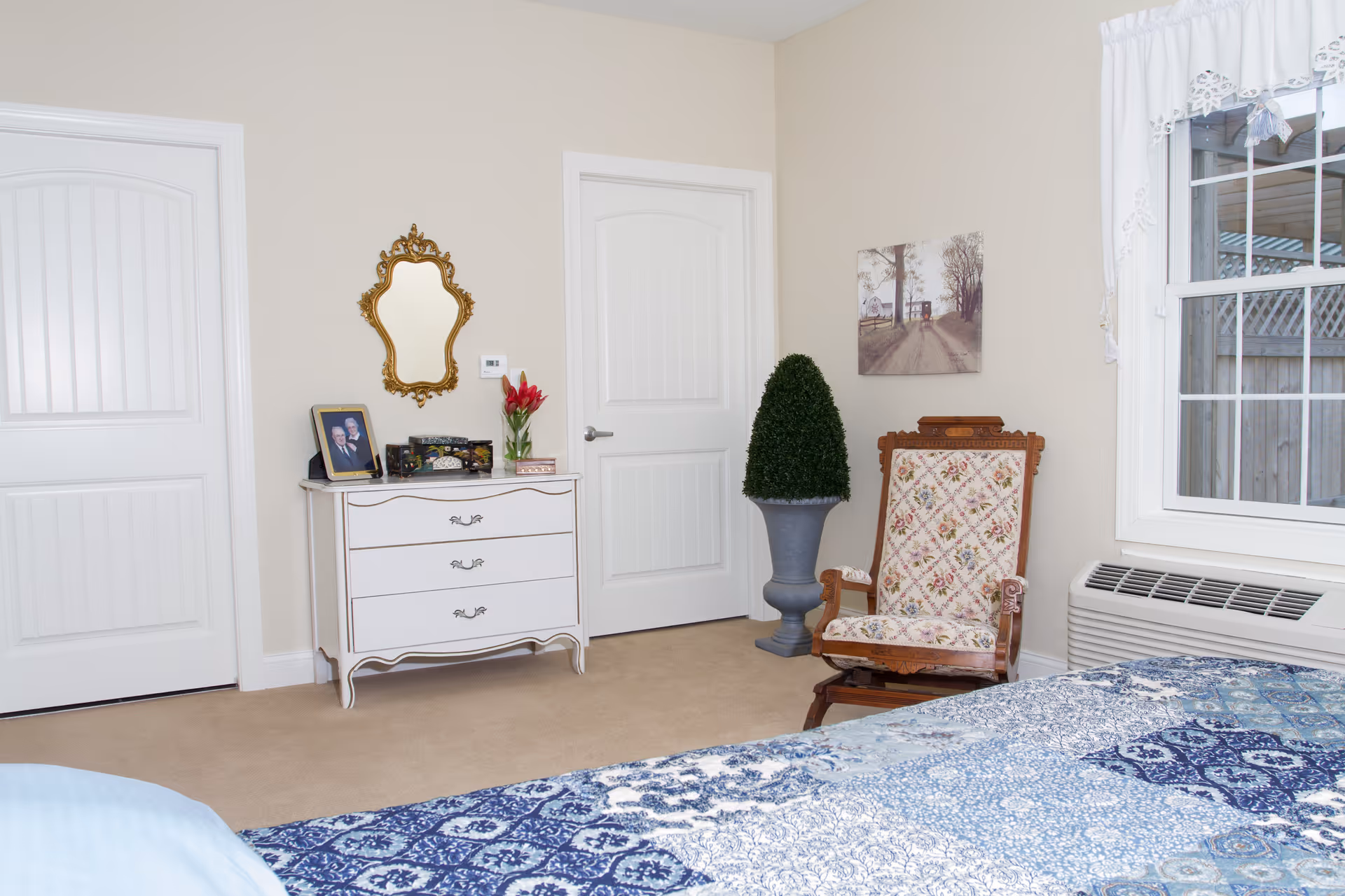 Sunlit bedroom interior featuring a blue-patterned bedspread, white dresser with a decorative mirror, an ornate wooden chair, and a window.